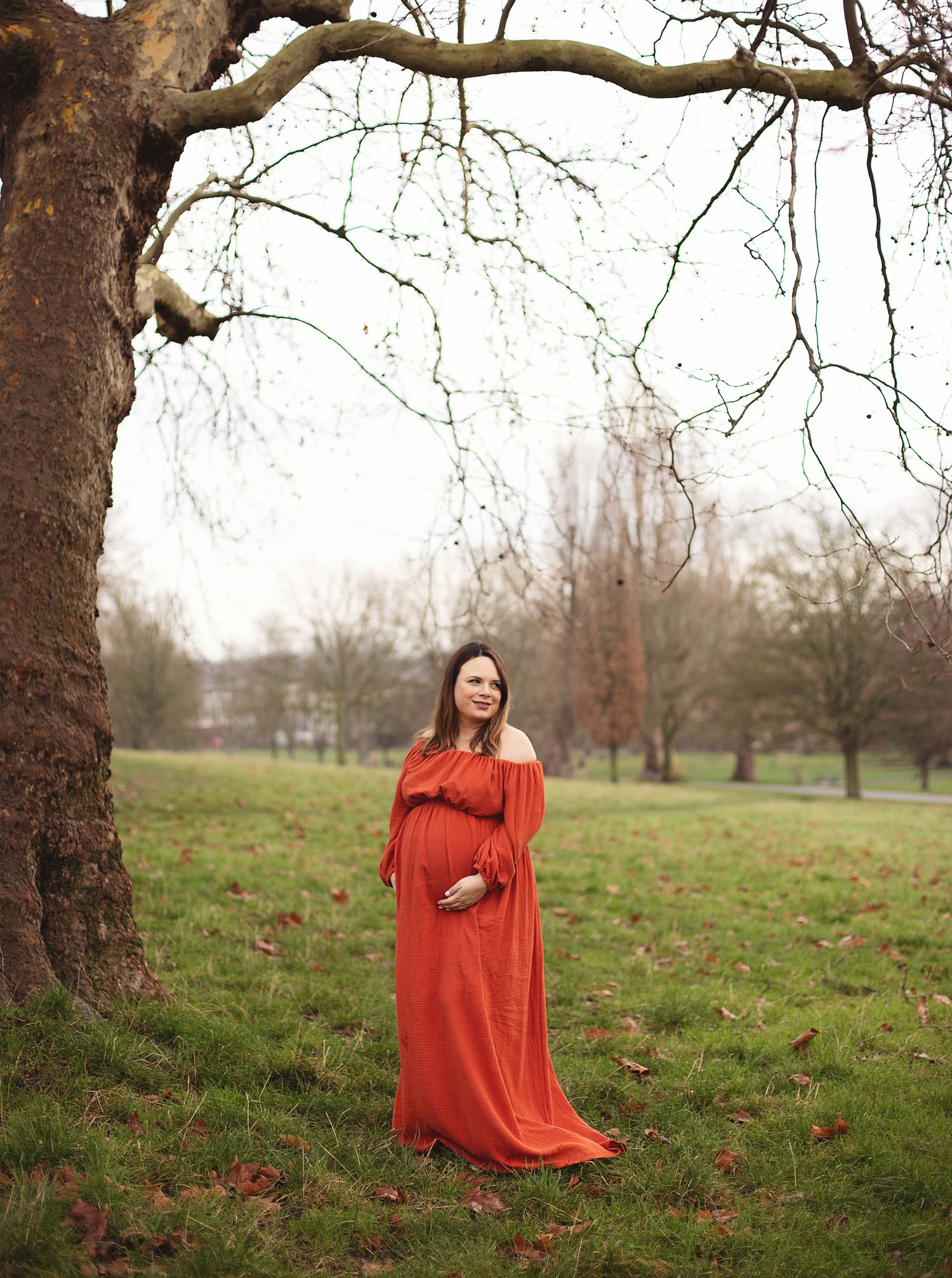 Pregnant woman in rust orange off-shoulder maxi dress standing under a tree in park during winter. during a maternity photoshoot in London