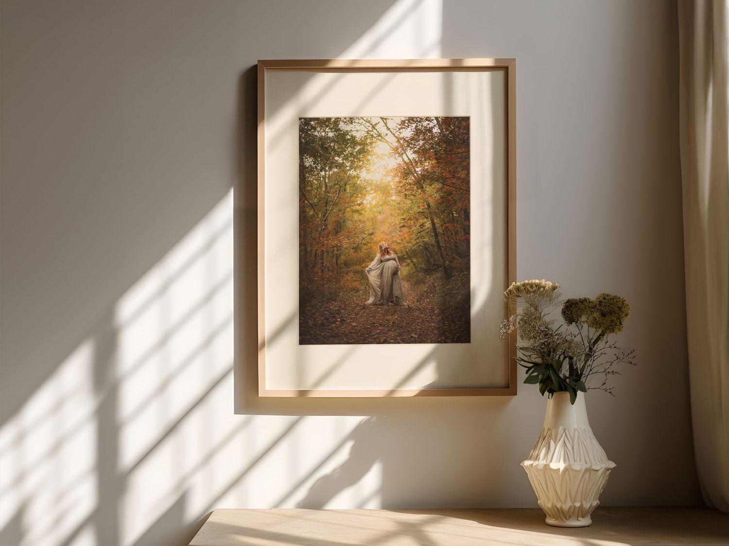 Framed autumn forest maternity photo print in wooden frame displayed on wall above a vase with dried flowers in warm sunlight.