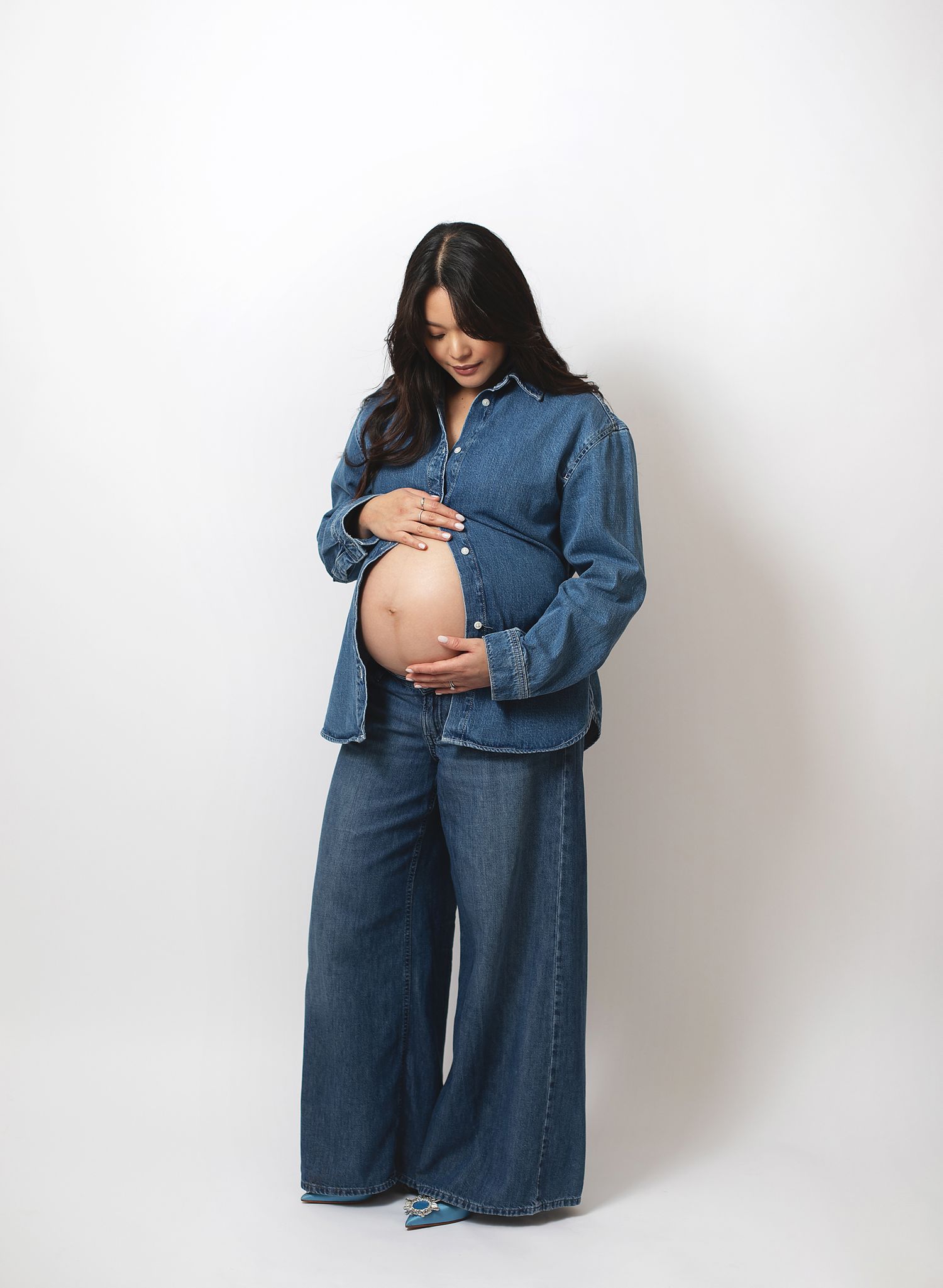 Pregnant woman in denim shirt and wide-leg jeans cradling baby bump, posing against white background. Photoshoot in London