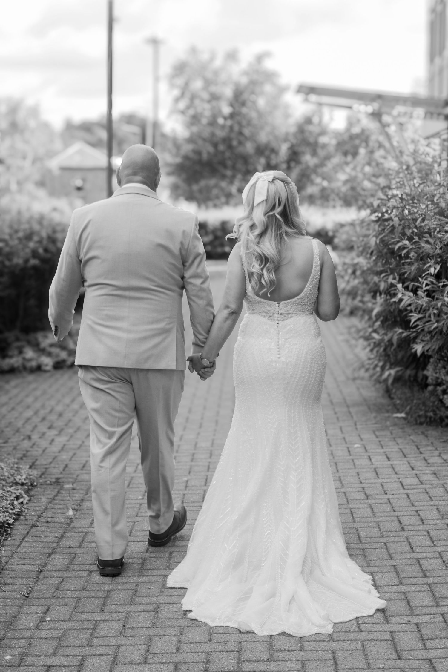 Black and white photo of a bride in a lace mermaid wedding gown holding hands with a groom as they walk down a brick path.