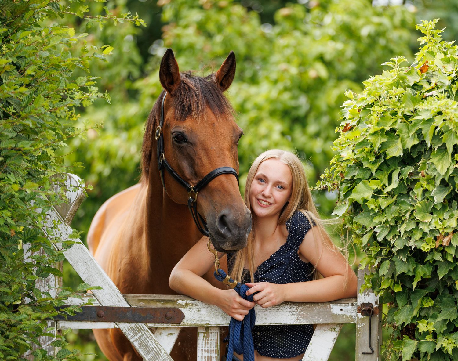 A woman sits on a log beside a brown horse in a field, holding its reins in a serene black and white photo.