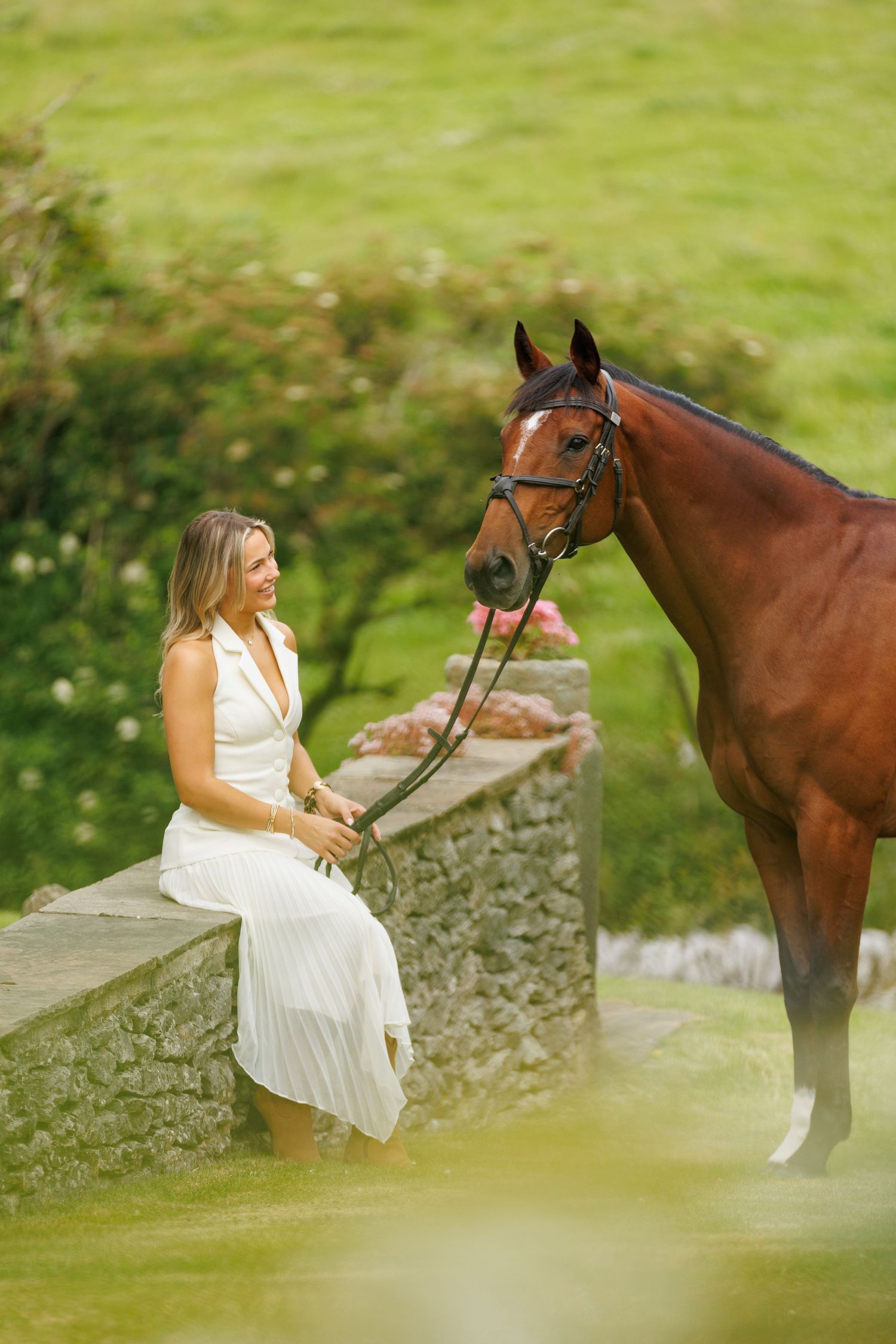 Young woman smiling between two bay horses with white markings, holding reins near a brick stable.