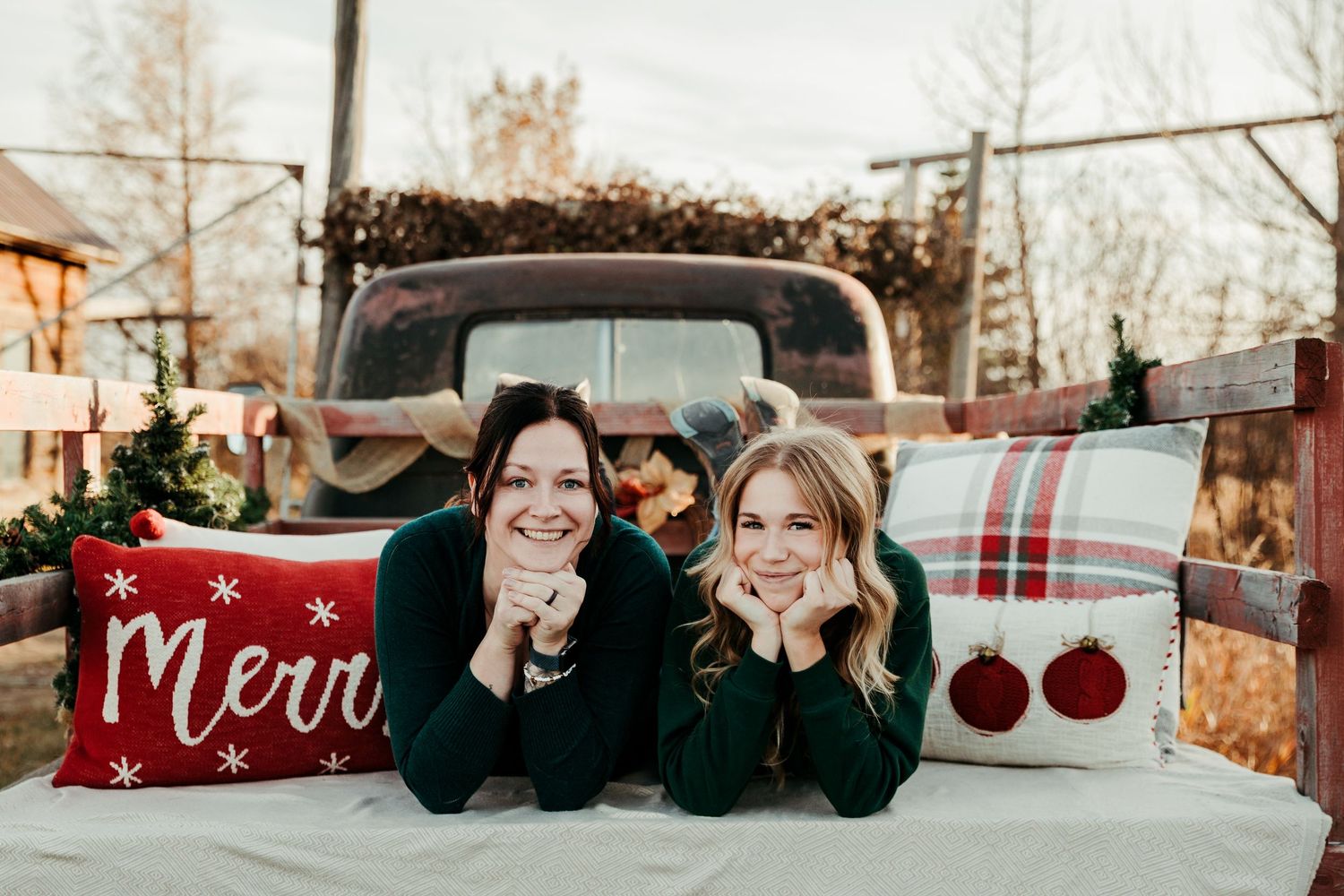 Two smiling women in green sweaters lie on a truck bed with festive red Merry Christmas pillows in front of a vintage pickup.