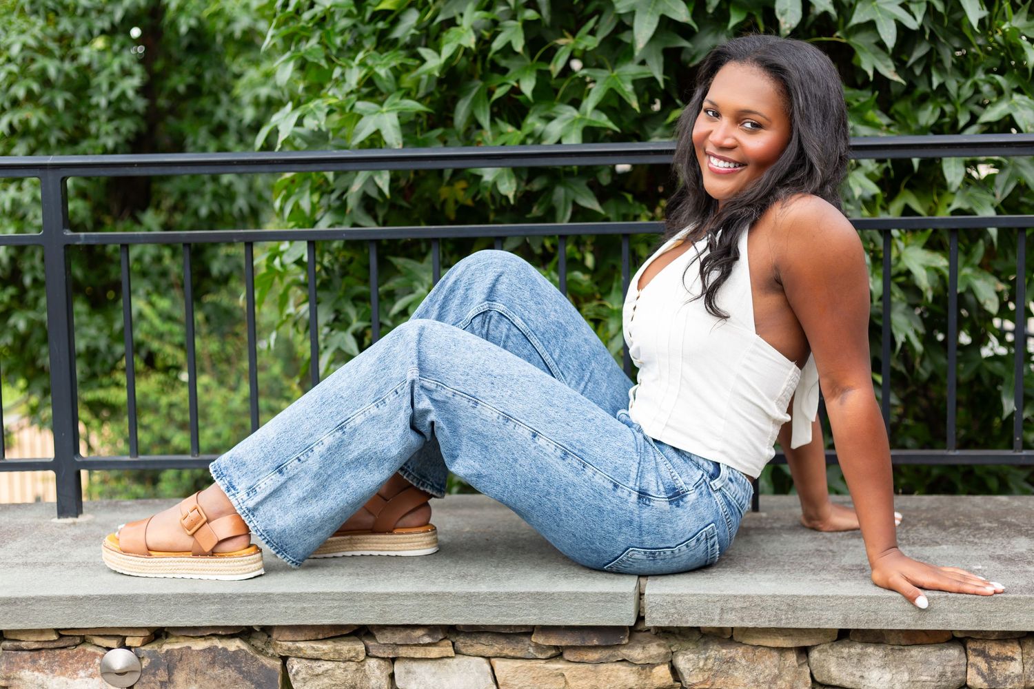 Smiling woman in a white halter top and high-waisted jeans walks along a sandy garden path beside lush green foliage.
