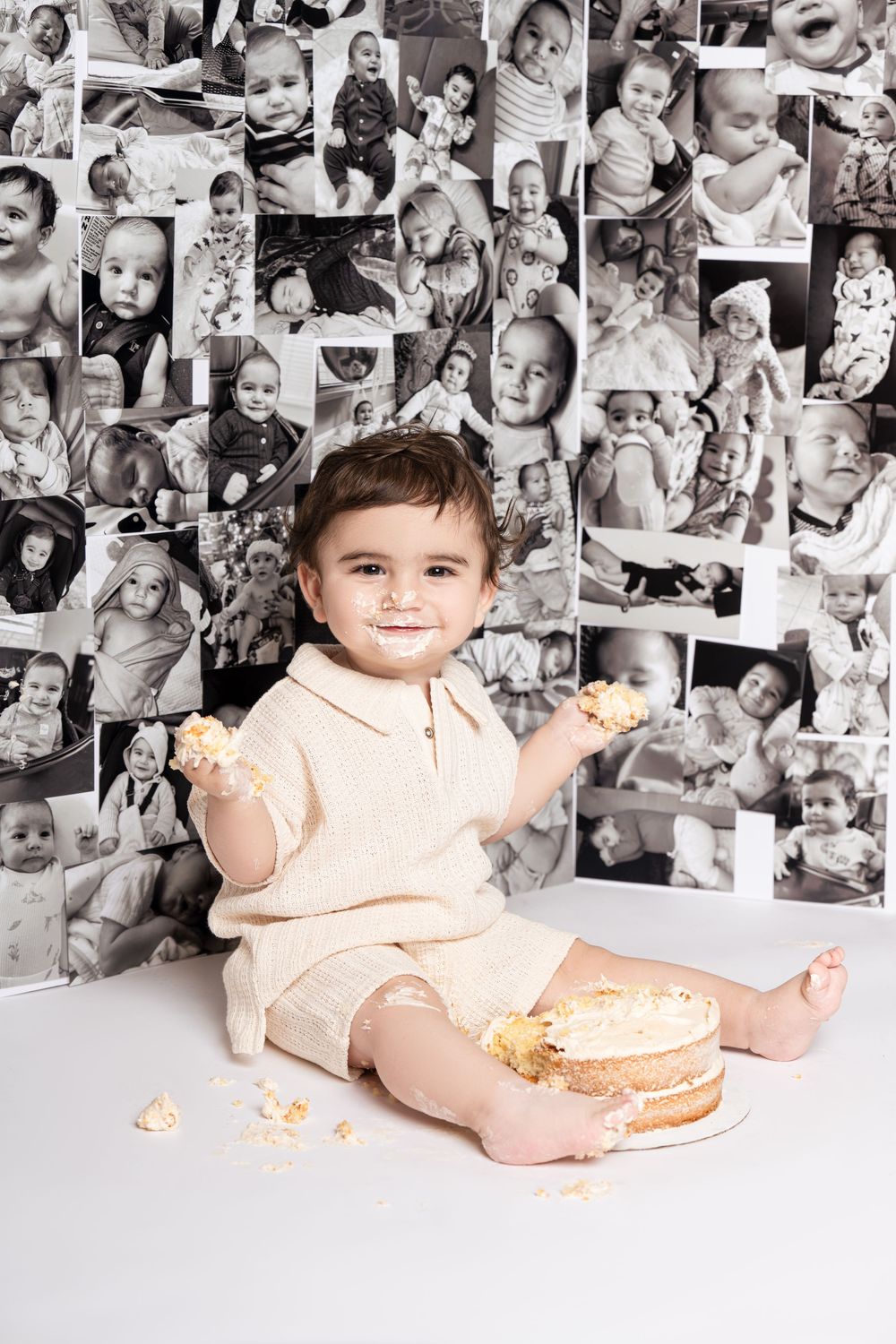 Smiling toddler sitting beside a birthday cake with a black-and-white photo collage backdrop.