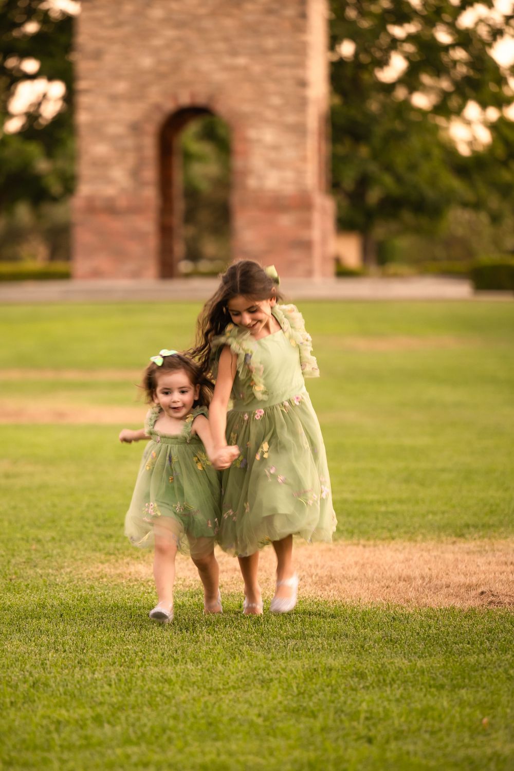 Sisters in matching green dresses walking together across a sunlit lawn.