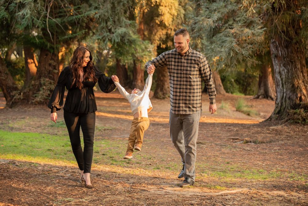 Parents playfully swinging their young child by the hands while walking along a tree-lined path.