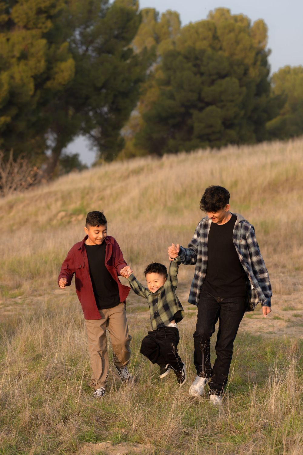 Three children walking together through tall grass on a hillside during an outdoor family photoshoot.