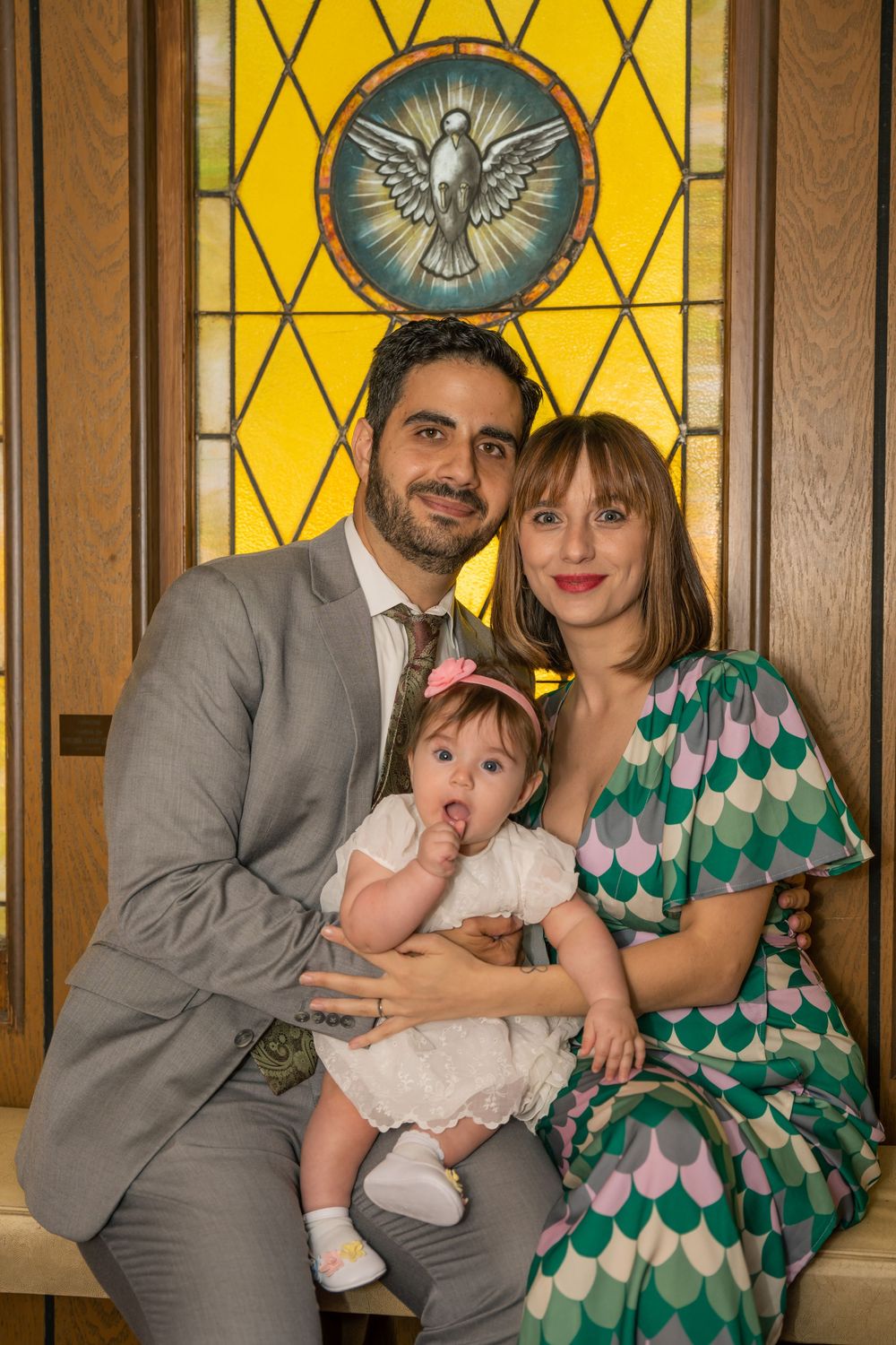Parents posing with their baby during a Christian christening celebration in front of a stained-glass window.