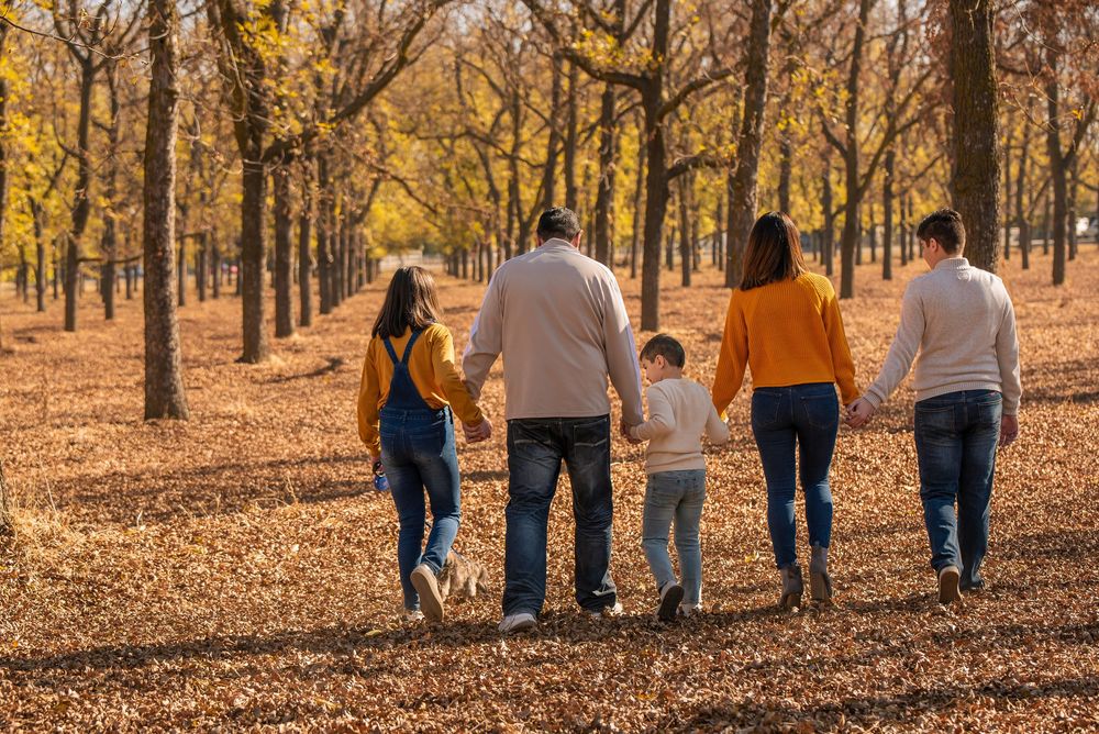 Family of five walking hand in hand through a tree-lined path covered in golden autumn leaves.
