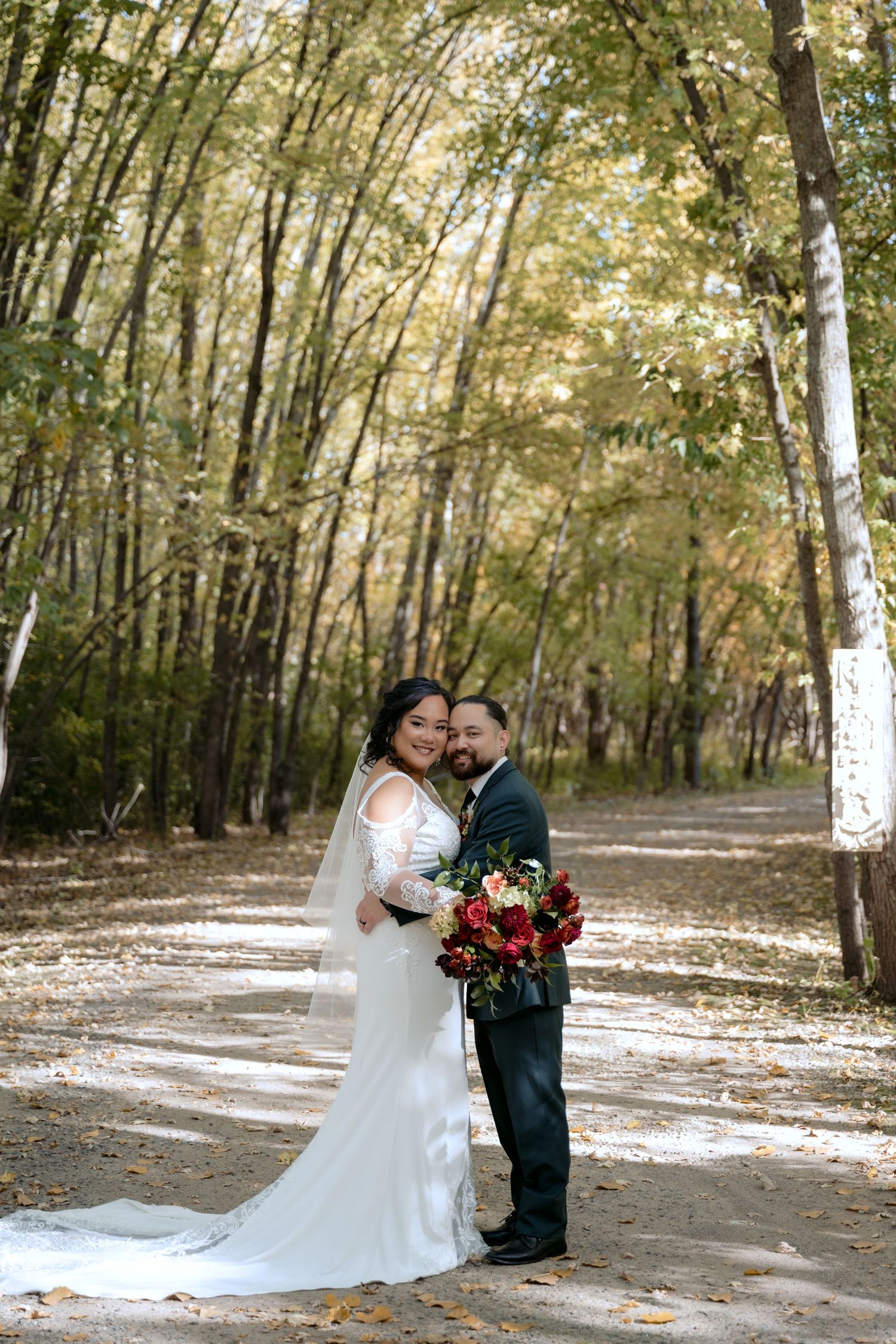 Bride and Groom stand together on a path surrounded by tall trees and fall colors at Trellis Weddings in Stillwater, MN.