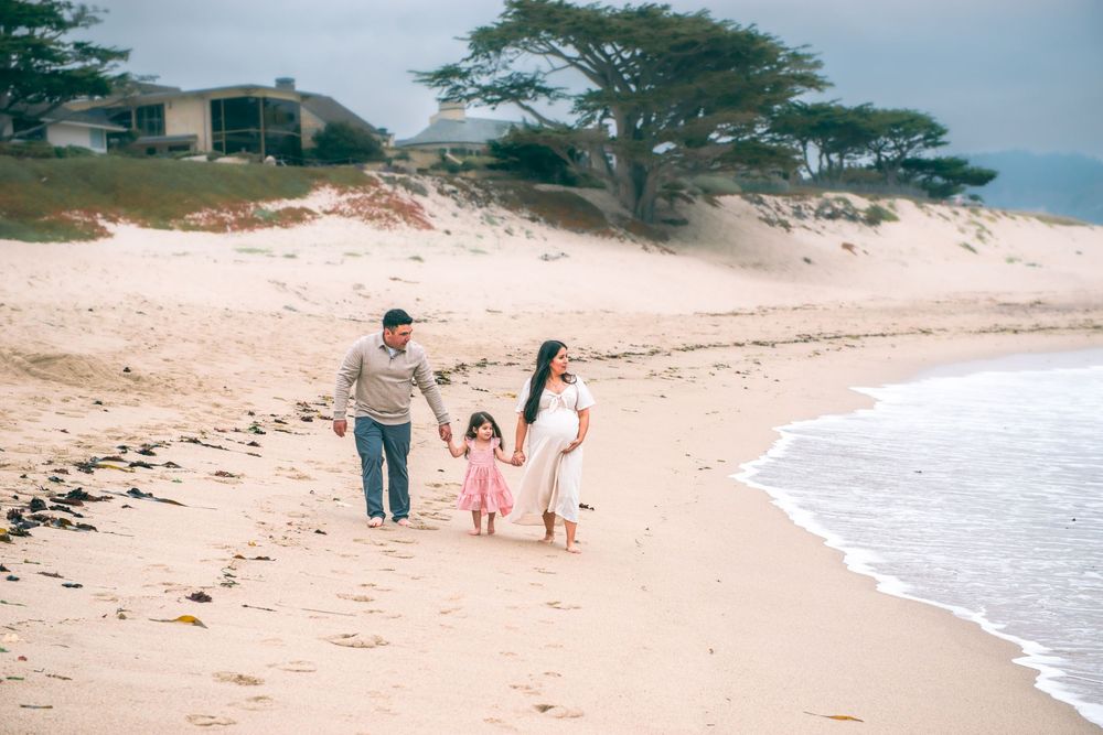 Family of three holding hands while walking along a sandy beach near the shoreline, with coastal trees and a house in the background.