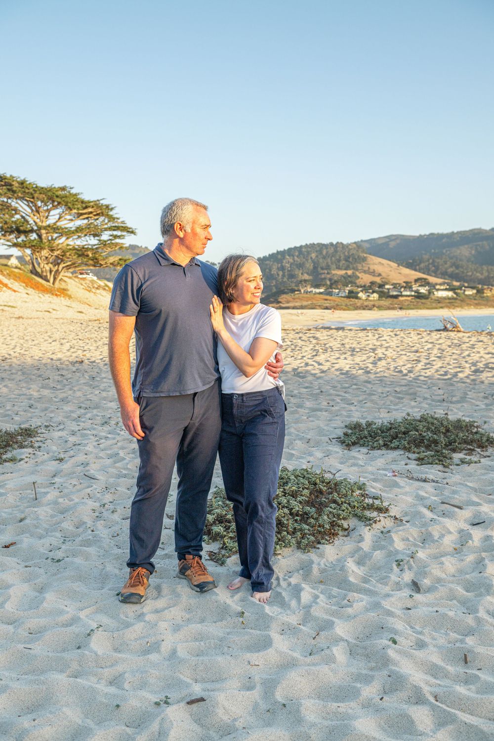 Smiling couple embracing on a sandy beach at sunset with a cypress tree, calm bay, and coastal hills in the background.