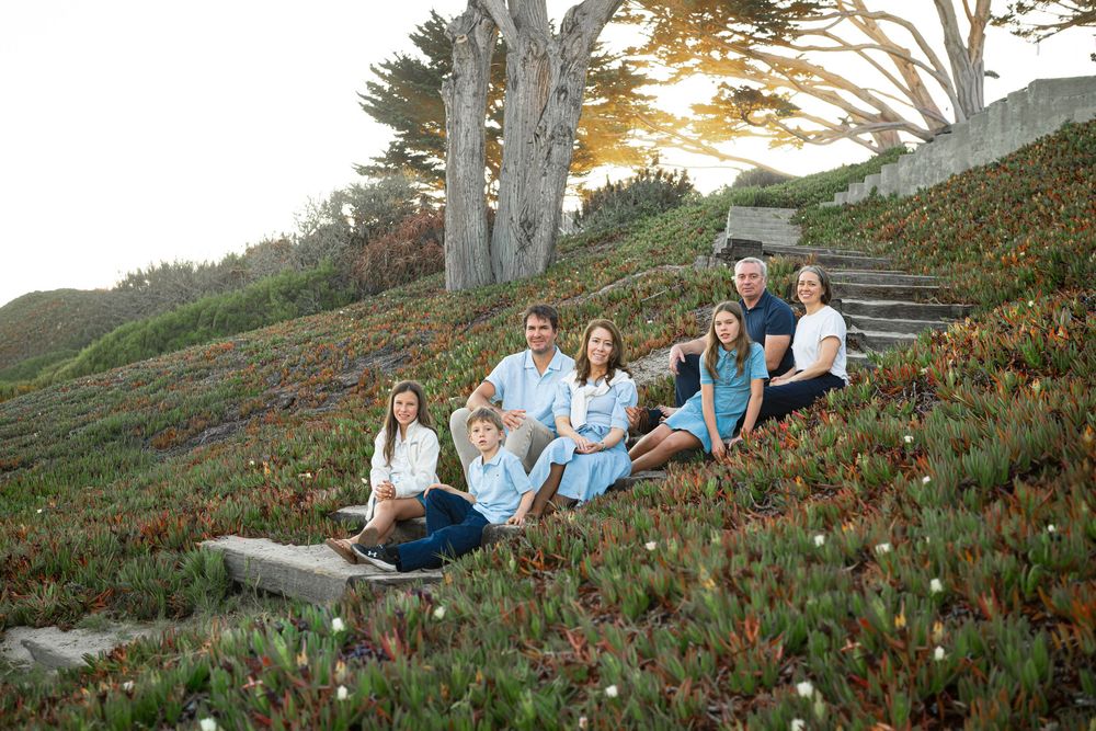 Family of seven in blue and white outfits sits together on stone steps surrounded by green ice plants at golden hour near cypress trees.