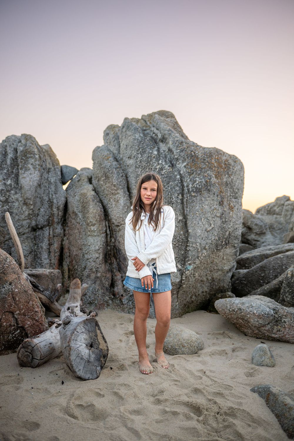 A young girl in a white jacket and denim shorts stands barefoot on a sandy beach beside large granite boulders and driftwood at sunset.