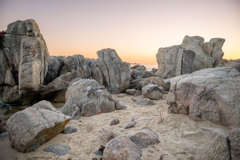 Large granite boulders scattered on sandy beach at sunset with warm orange and pink sky in background.