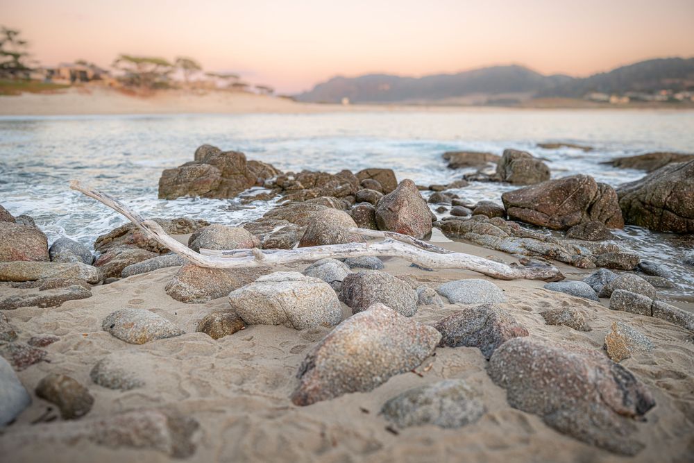 Rocky beach shoreline at sunset with smooth stones, gentle waves, and misty mountains in the background.