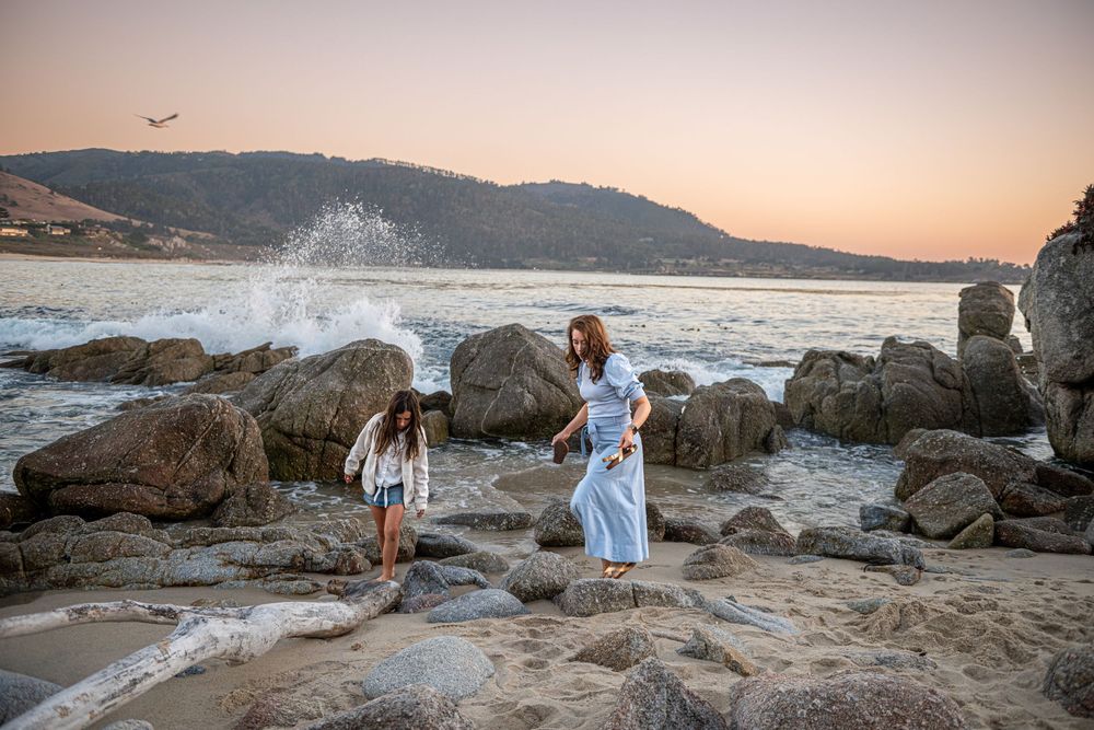 Woman in a light blue dress and a young girl walk among rocks on a sandy beach at sunset as ocean waves crash nearby.