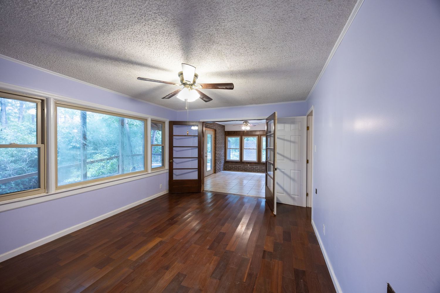 Empty room with hardwood floors, lavender walls, ceiling fan, and large windows letting in natural light.