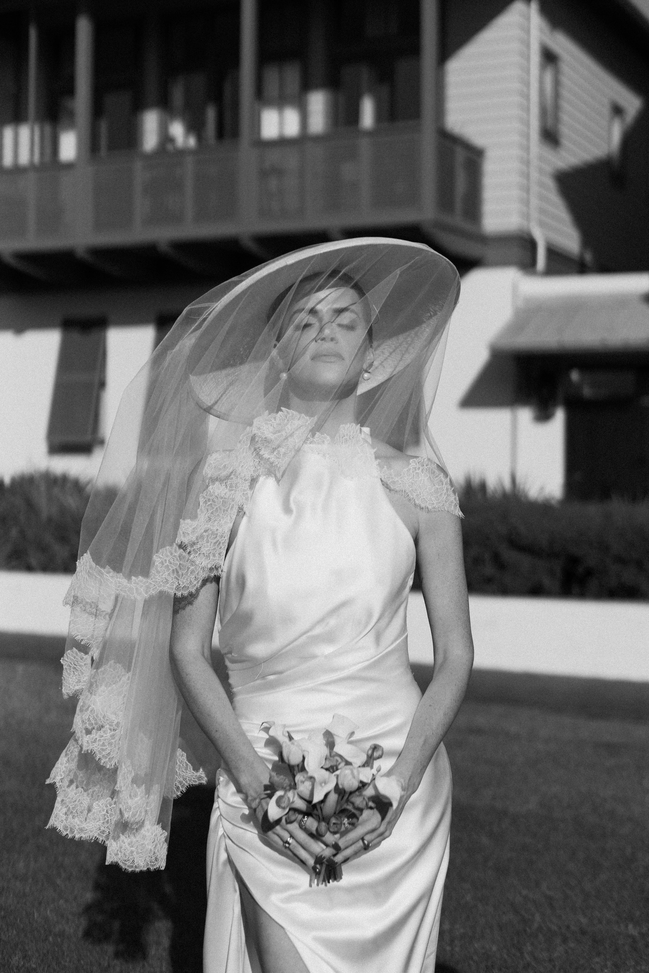 Black and white bridal portrait of a woman in satin gown with wide brim veil and bouquet.