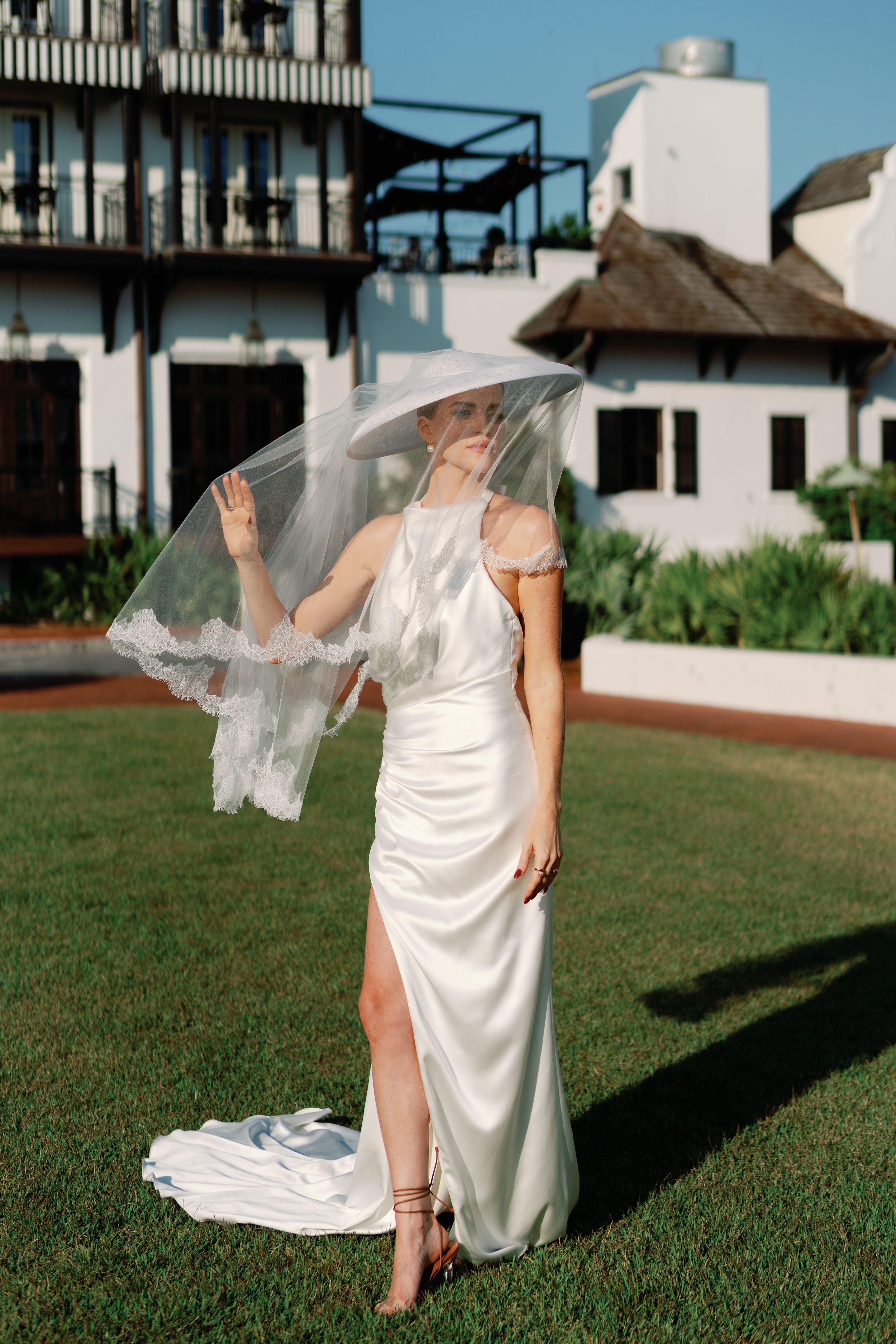 Bride in sleek white satin gown lifting lace-trimmed veil in the wind on a green lawn.