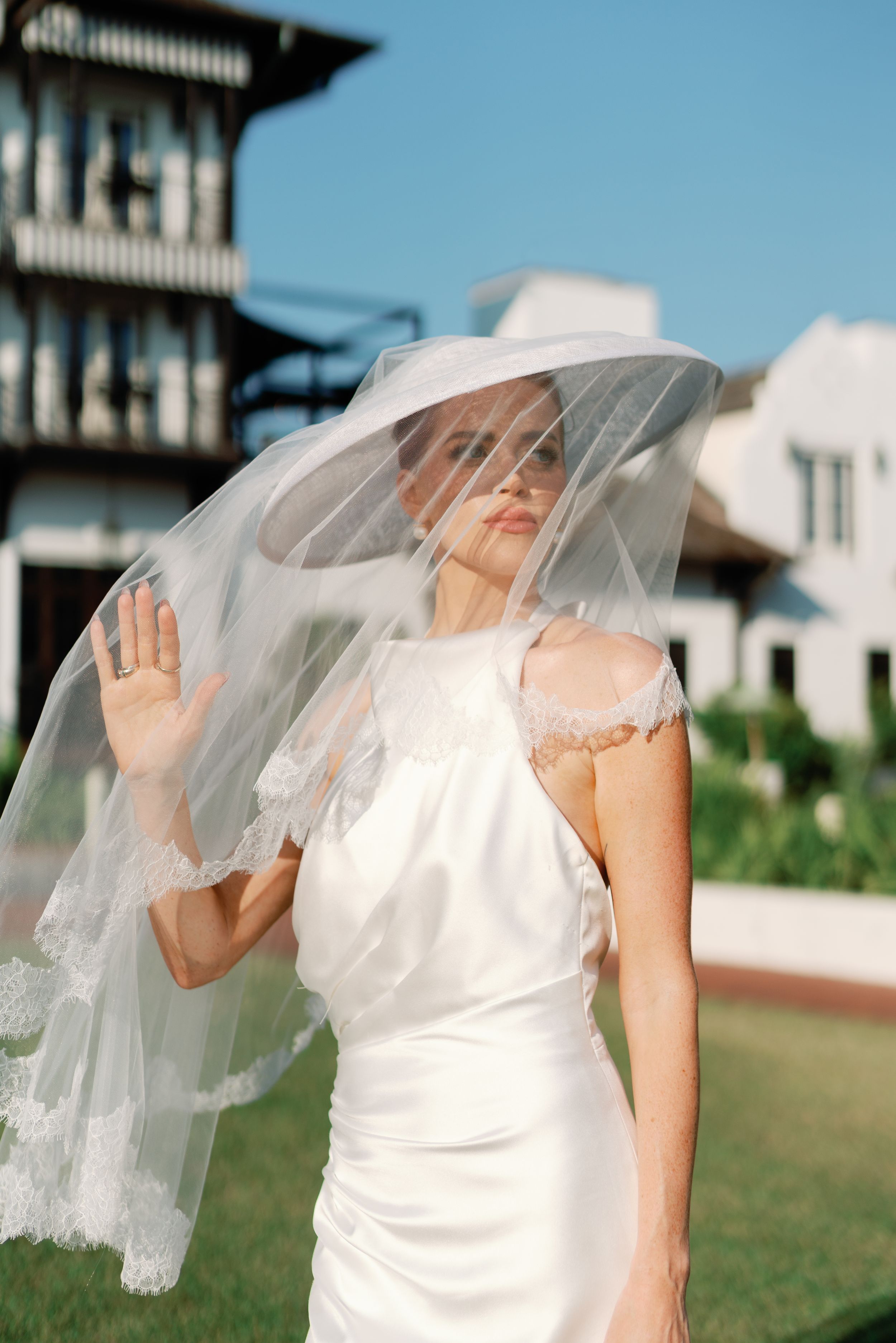 Bride in white satin dress holding up lace-edged veil with one hand against a blue sky.