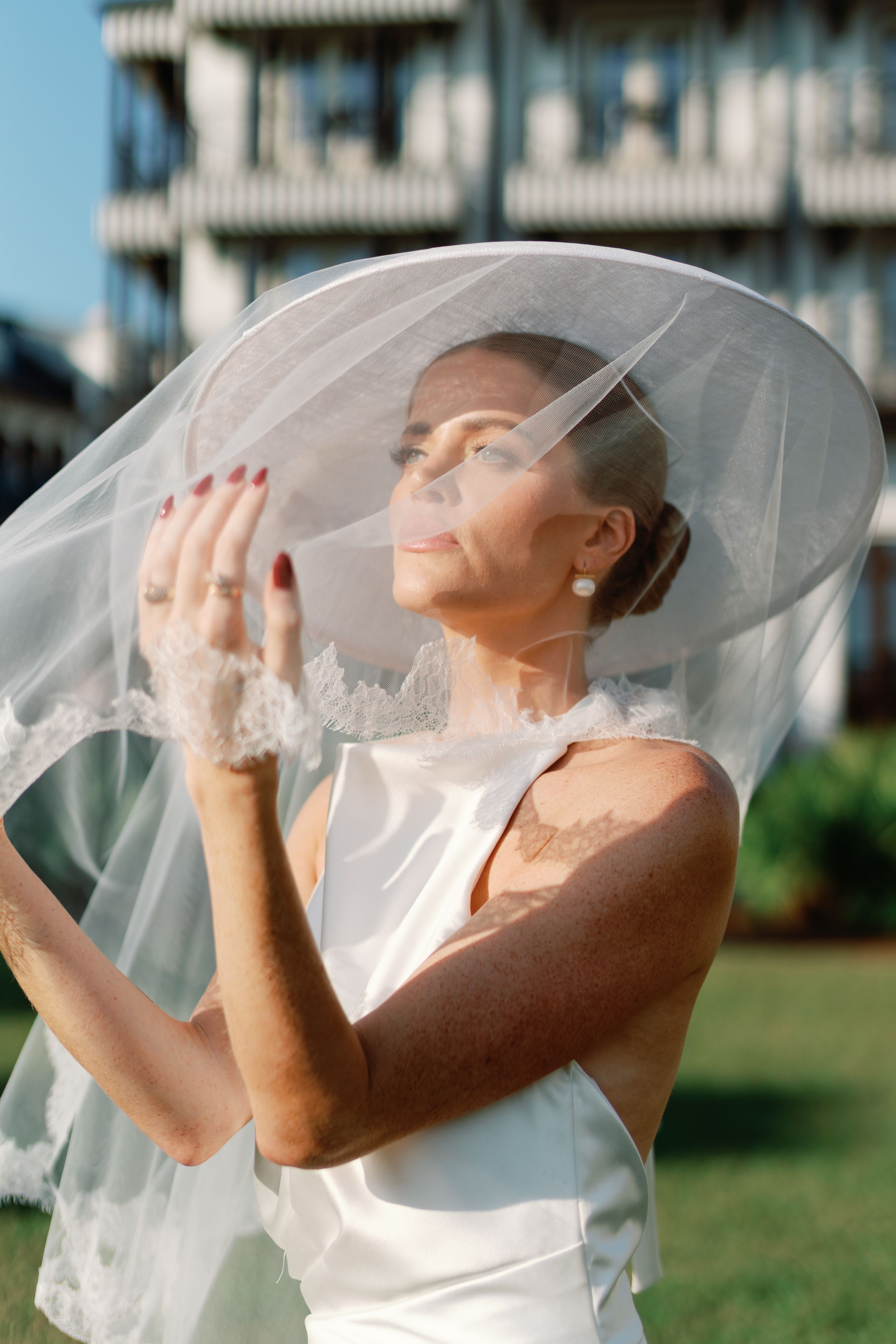 Bride in white satin gown lifting lace veil with both hands, eyes closed, outdoors.