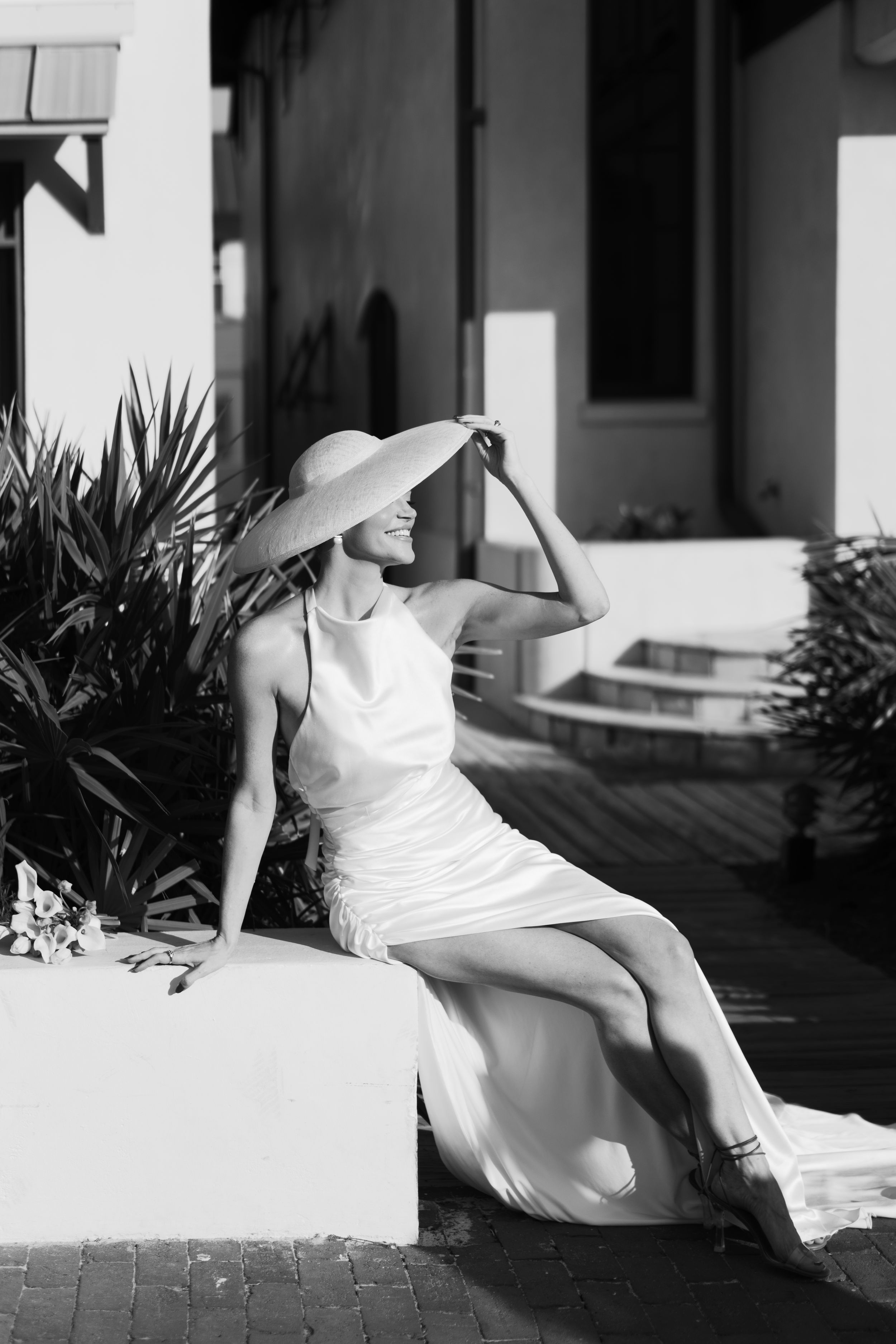 Bride in white silk gown and wide-brim hat sitting outdoors in black and white photo.