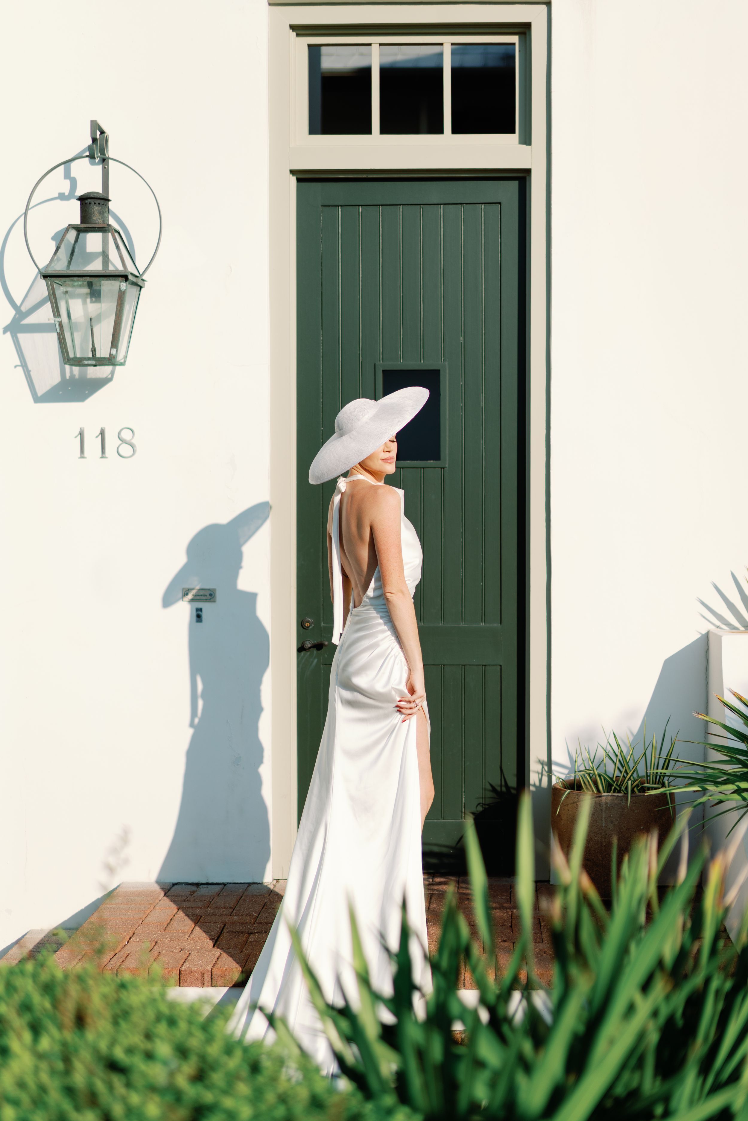 Bride in backless white gown and wide-brim hat standing before green door number 118.