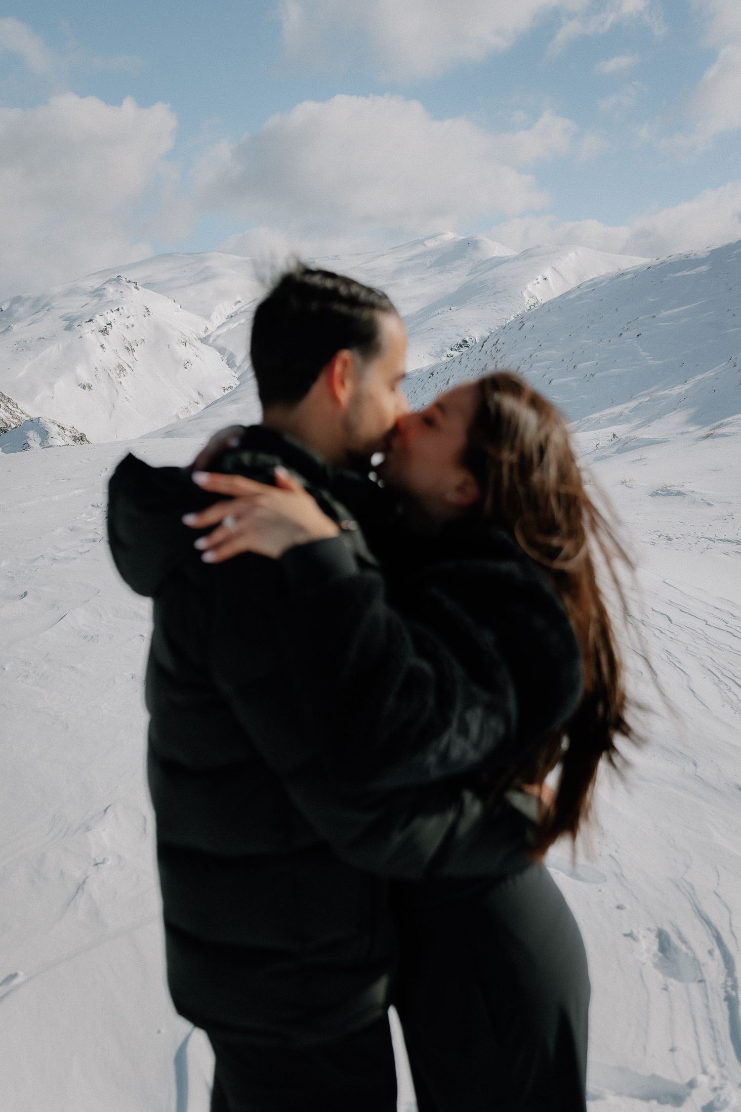 Couple sharing a romantic kiss on a snowy mountain slope, both wearing dark winter jackets with scenic alpine peaks behind them.