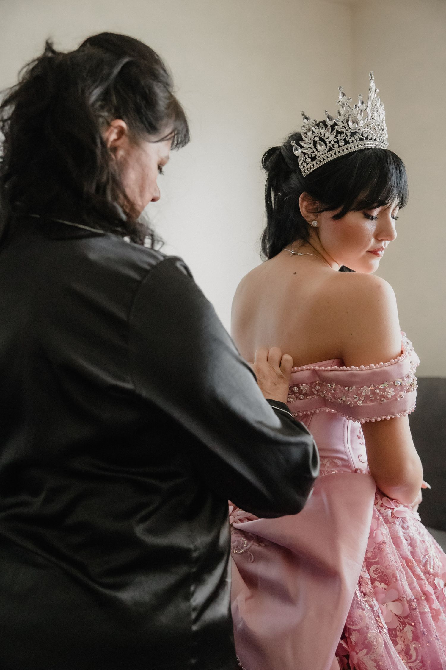 A woman in a black jacket helps a young girl in a pink off-shoulder gown and silver tiara prepare for her quinceañera.