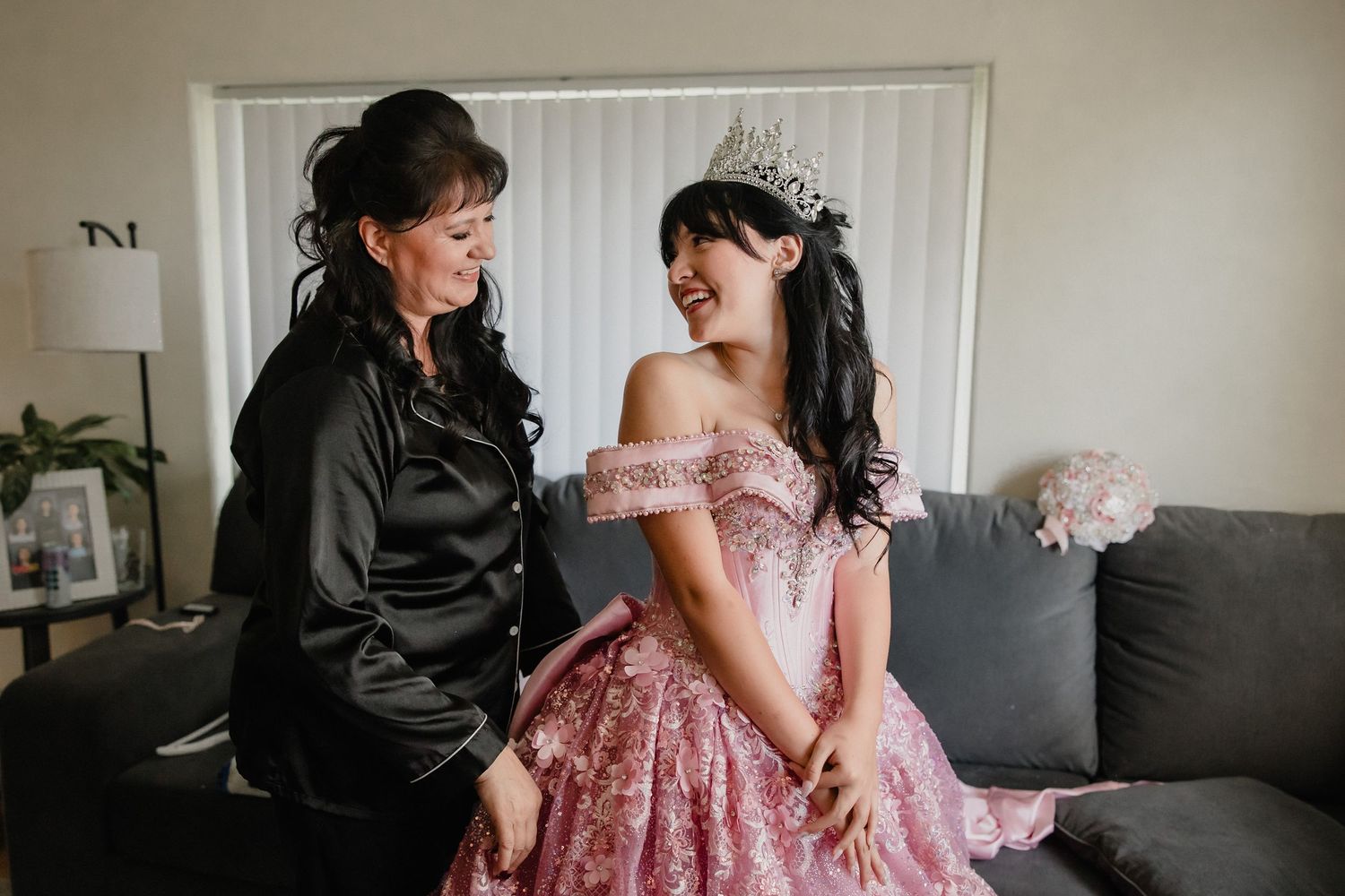 A young woman in a pink floral quinceañera gown and tiara smiles with an older woman in a black satin top.
