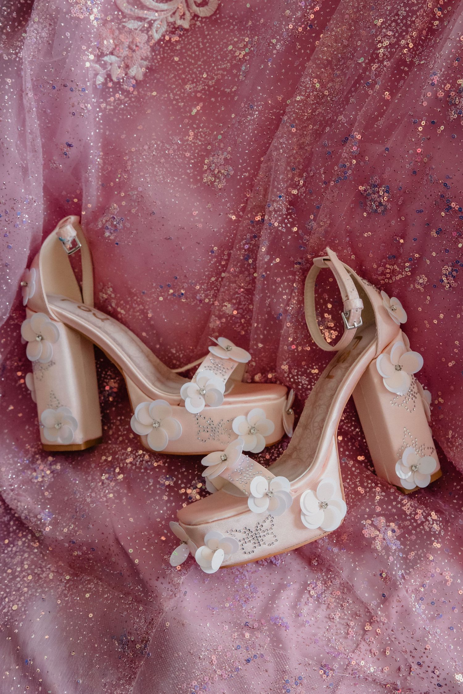 Elegant blush pink platform heels adorned with white floral appliques displayed on a sparkly rose pink fabric background.