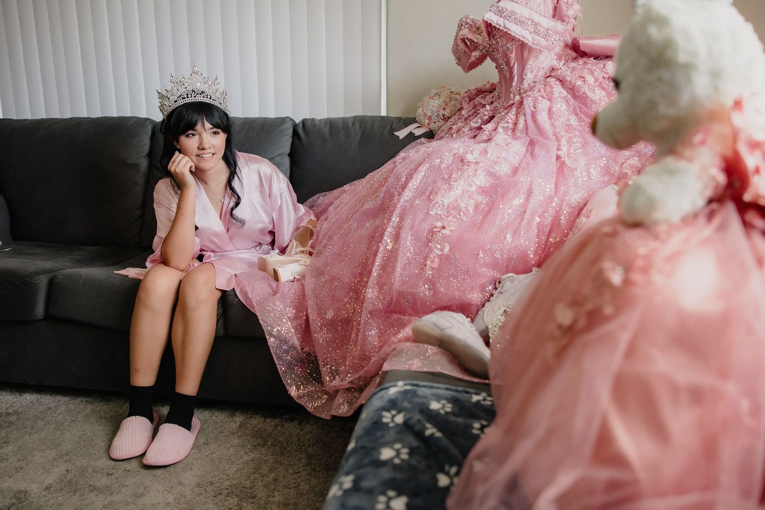 Young girl in pink robe and tiara sits on sofa beside an elegant pink quinceañera ball gown on a dress form.