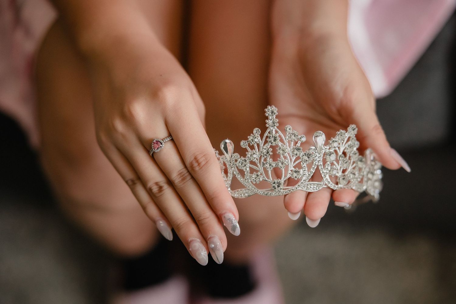 Close-up of elegant hands holding a sparkling silver tiara crown with crystal embellishments and white manicured nails.