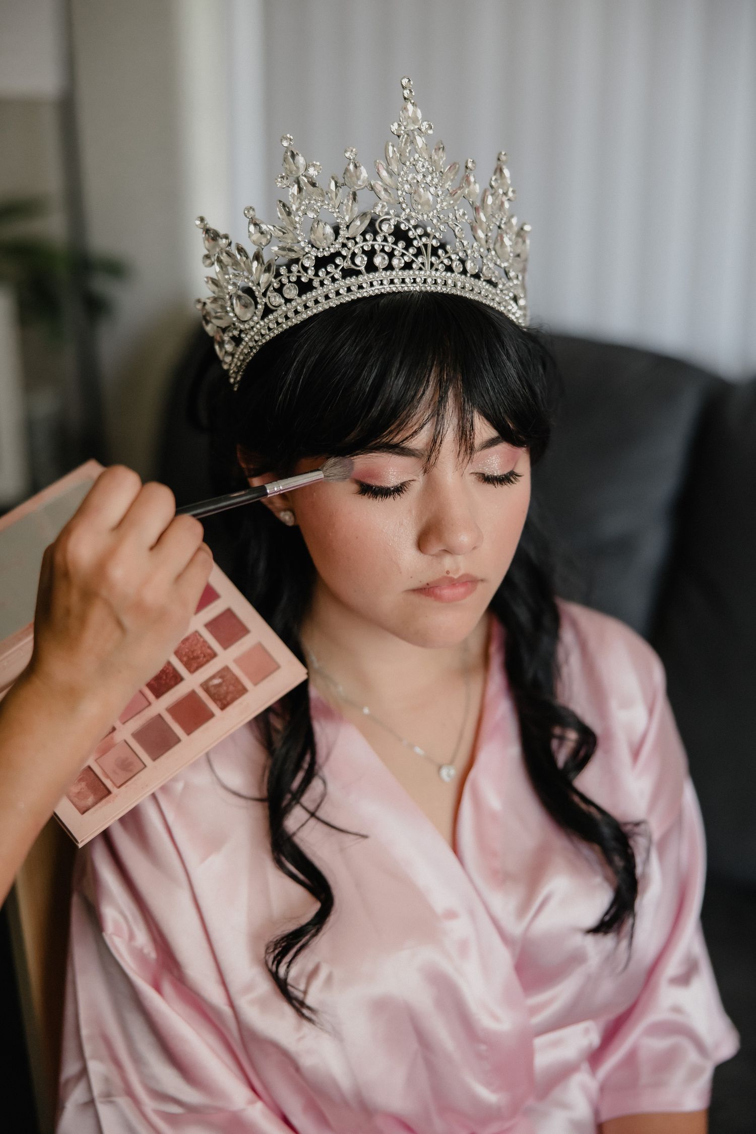 A bride wearing a silver crystal crown gets makeup applied while dressed in a pink satin robe.