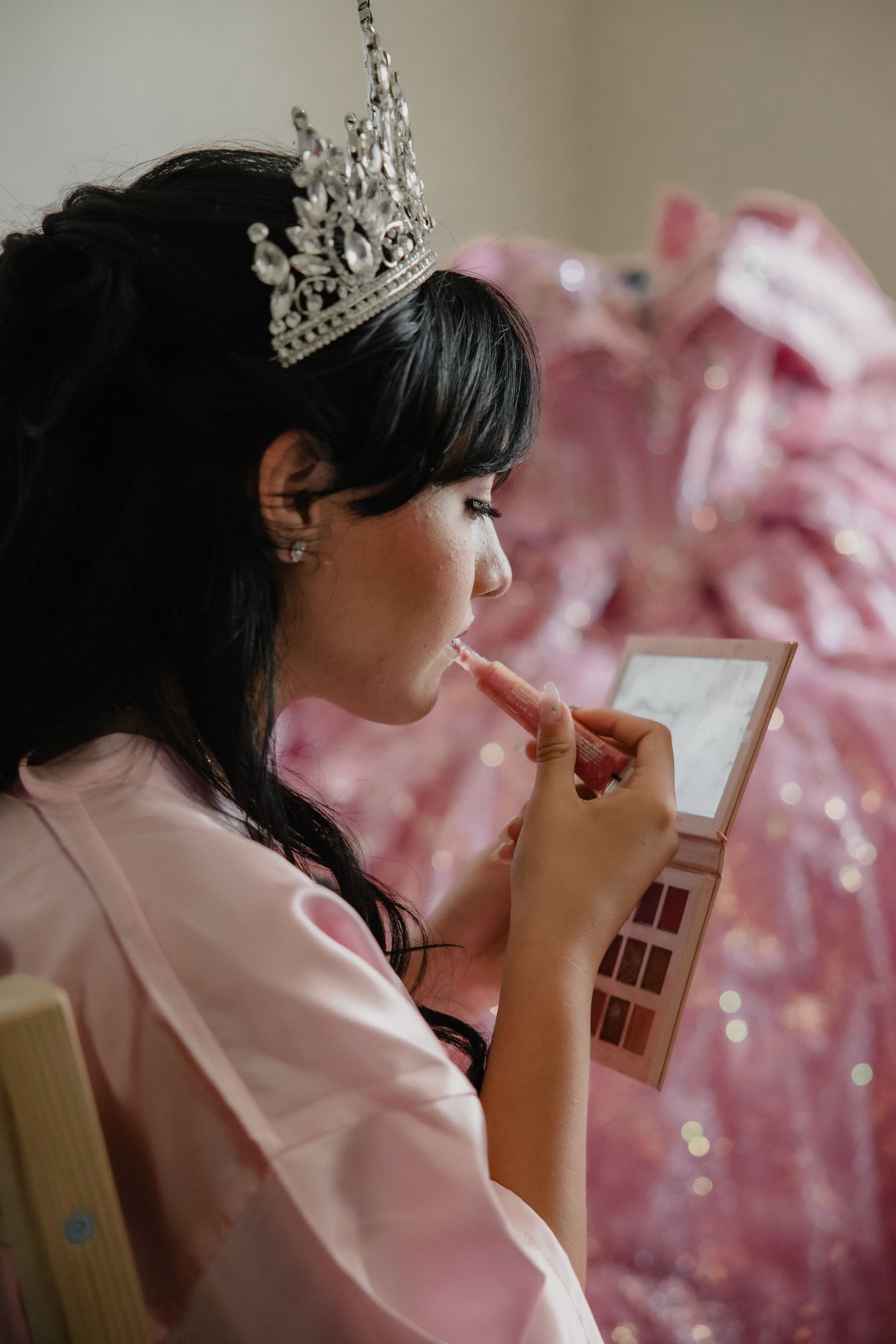A young woman in a pink robe and silver tiara applies lipstick using a compact mirror during quinceañera preparations.