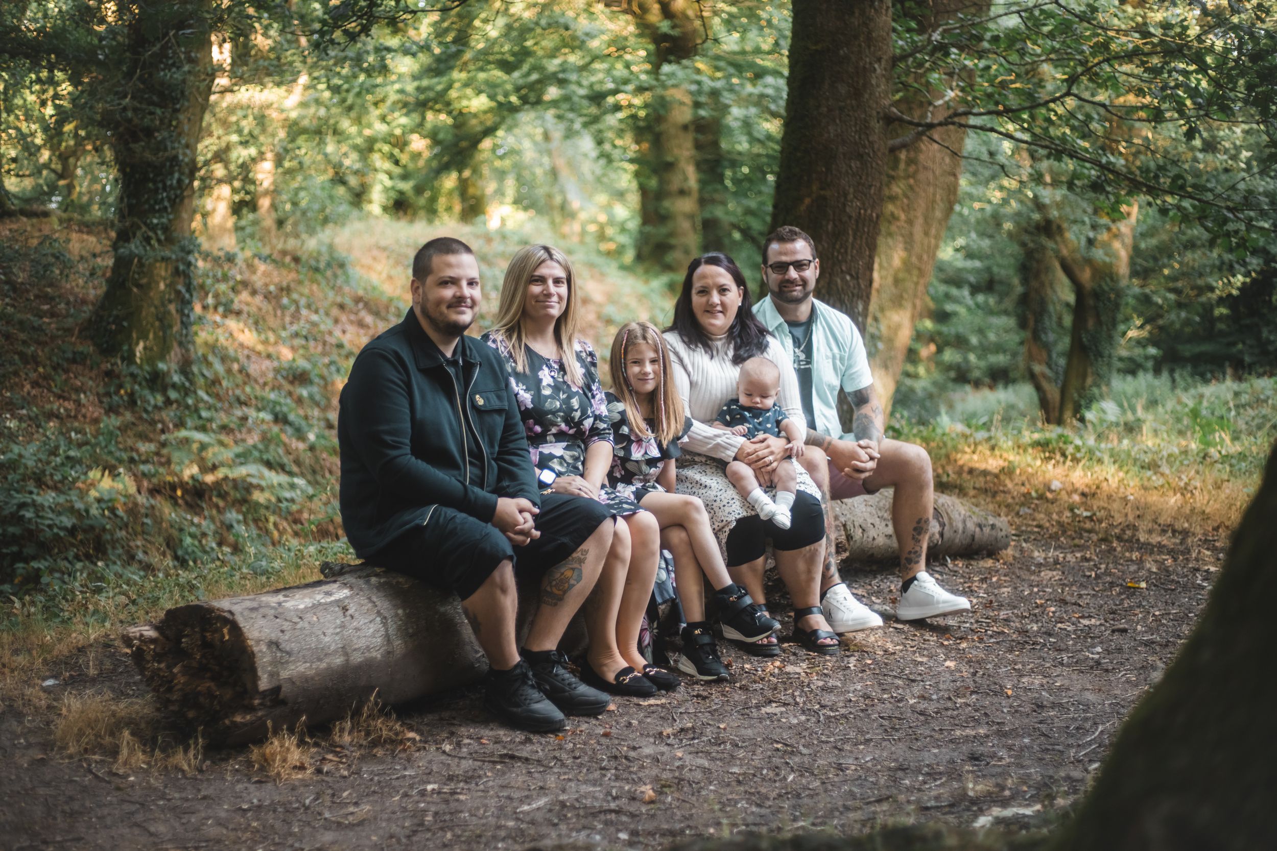 Family of six sitting on a log in a lush woodland setting, smiling together for an outdoor portrait.