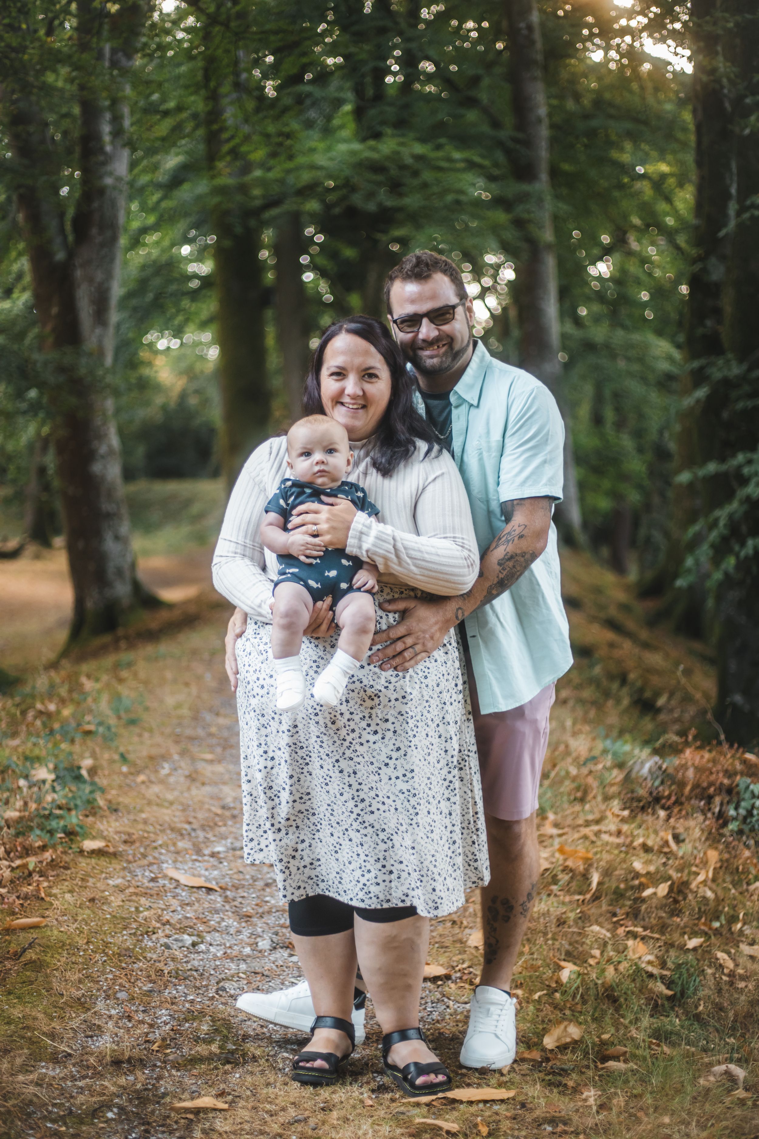 A happy family of three poses on a tree-lined path, with a woman holding a baby and a tattooed man beside her.