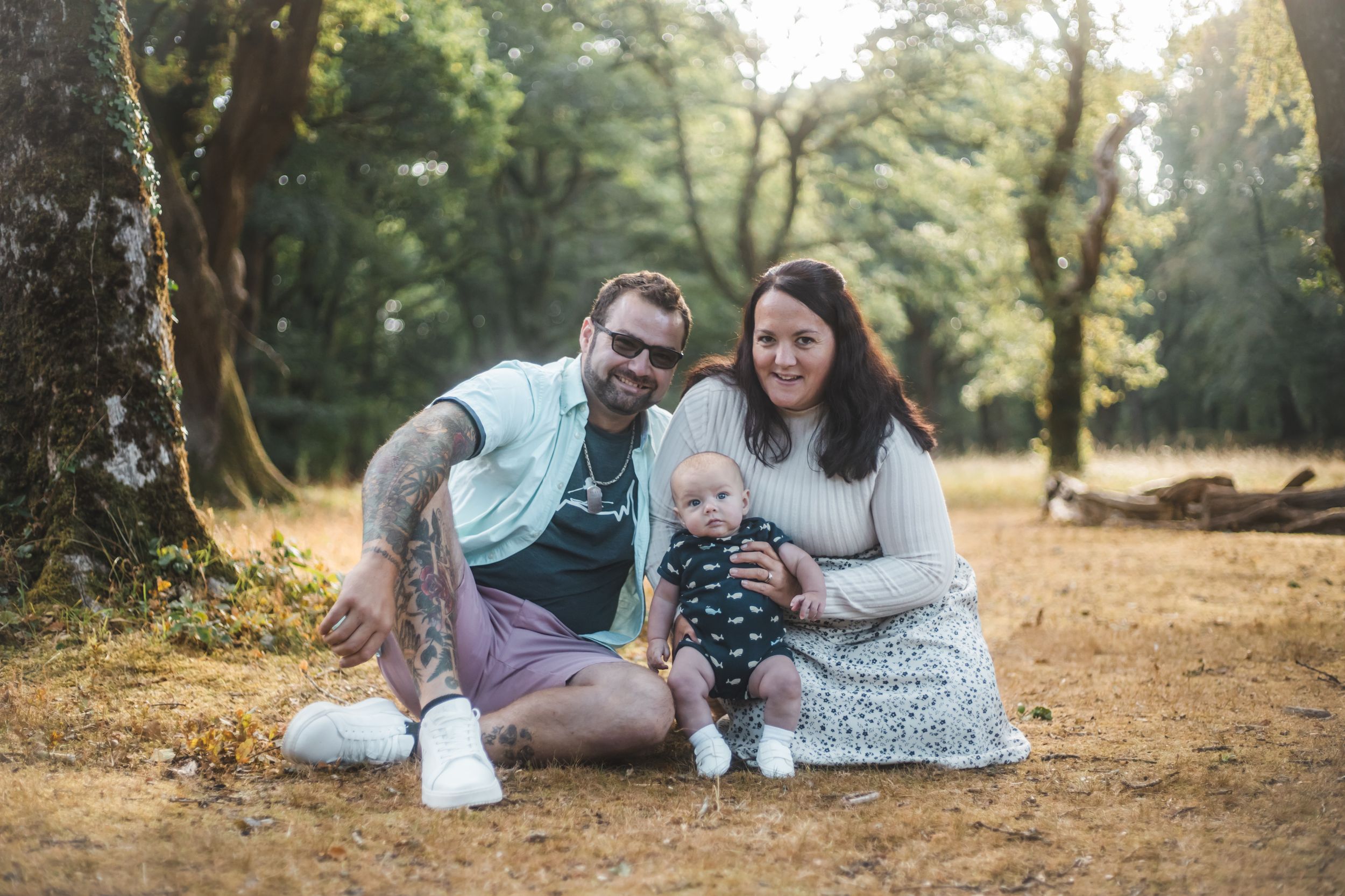 A tattooed man and woman sit outdoors in a forest park smiling with their baby in a striped onesie.