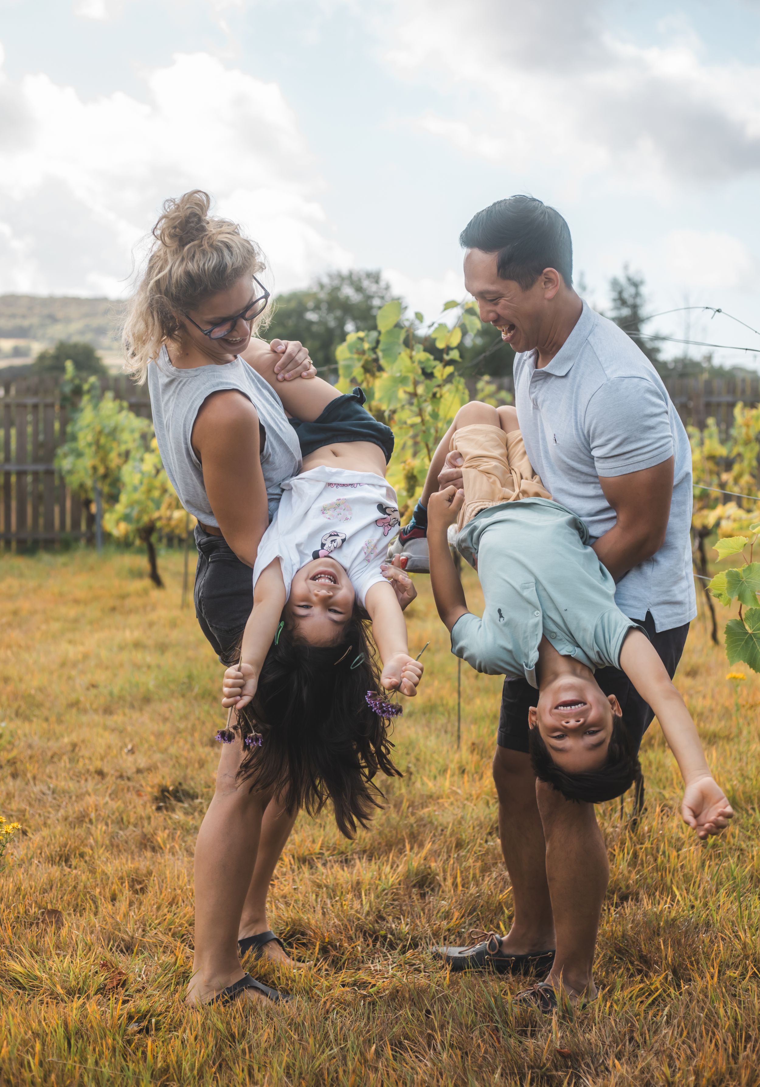 Family playfully holds two children upside down in a sunny vineyard, laughing and enjoying a fun outdoor moment together.