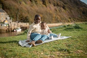 A young Black boy sits on a blanket outdoors with a baby, smiling, with a scenic hillside and water in the background.