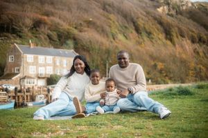 Happy Black family of four sitting on grass by a scenic coastal hillside with autumn foliage and a white building.