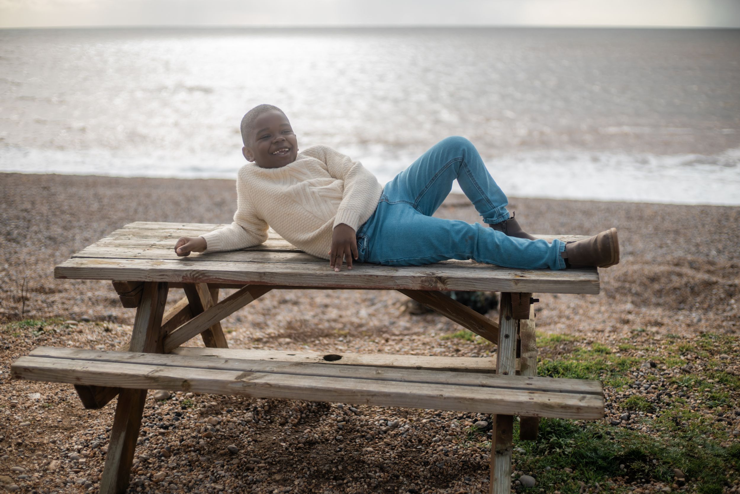 A man in a white sweater and blue jeans relaxes on a wooden picnic table on a pebble beach by the sea.