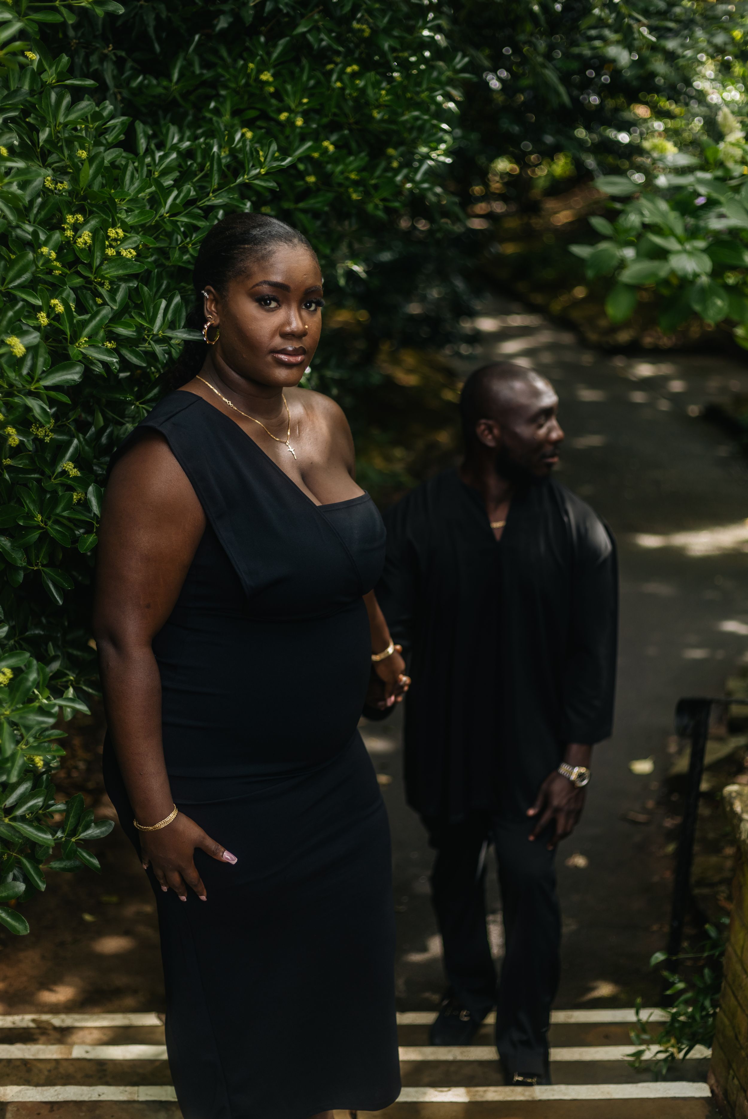 A couple in elegant black outfits poses on garden steps surrounded by lush green foliage.