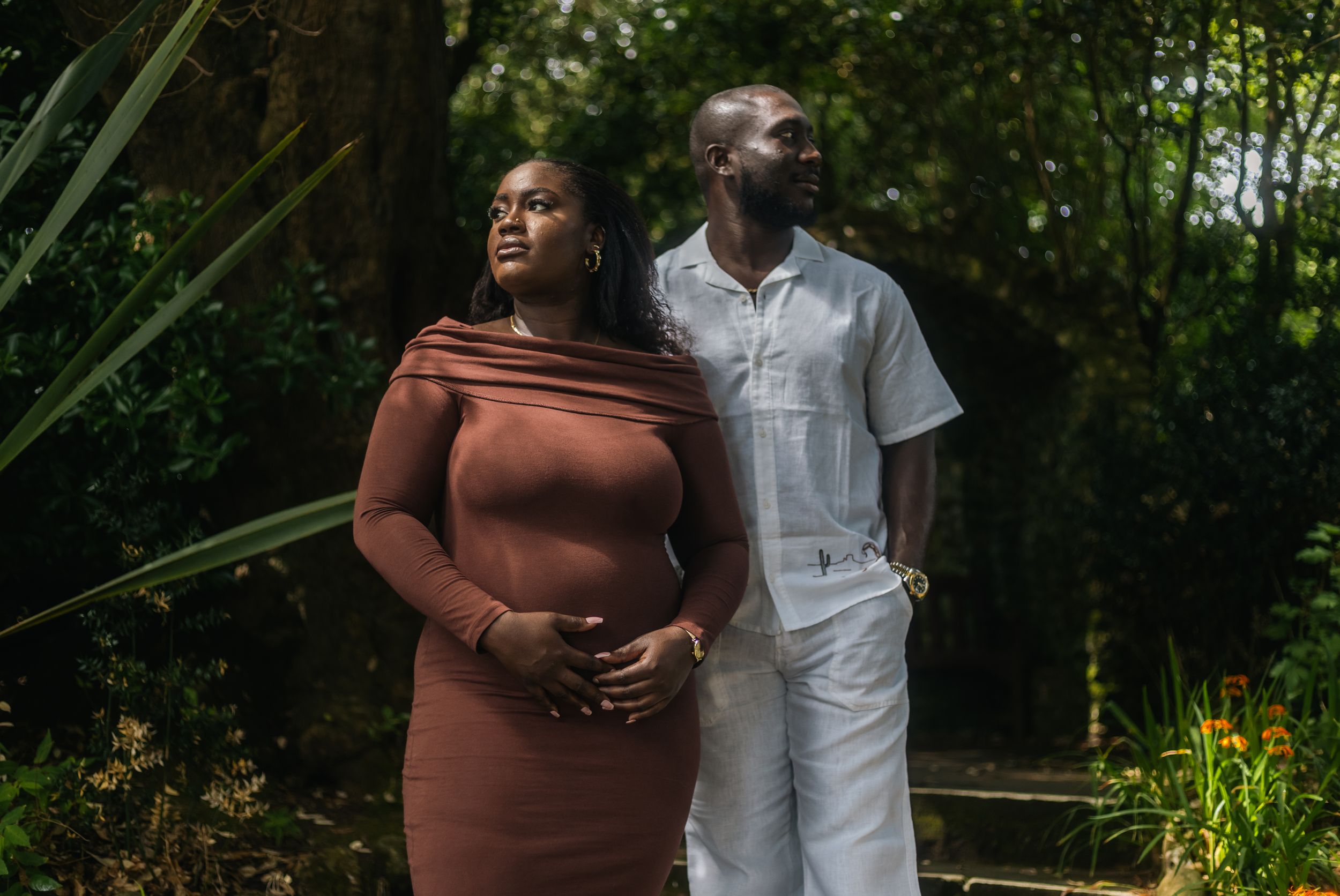 A pregnant Black couple poses in a lush garden, woman in brown dress, man in white outfit, sharing a tender moment.