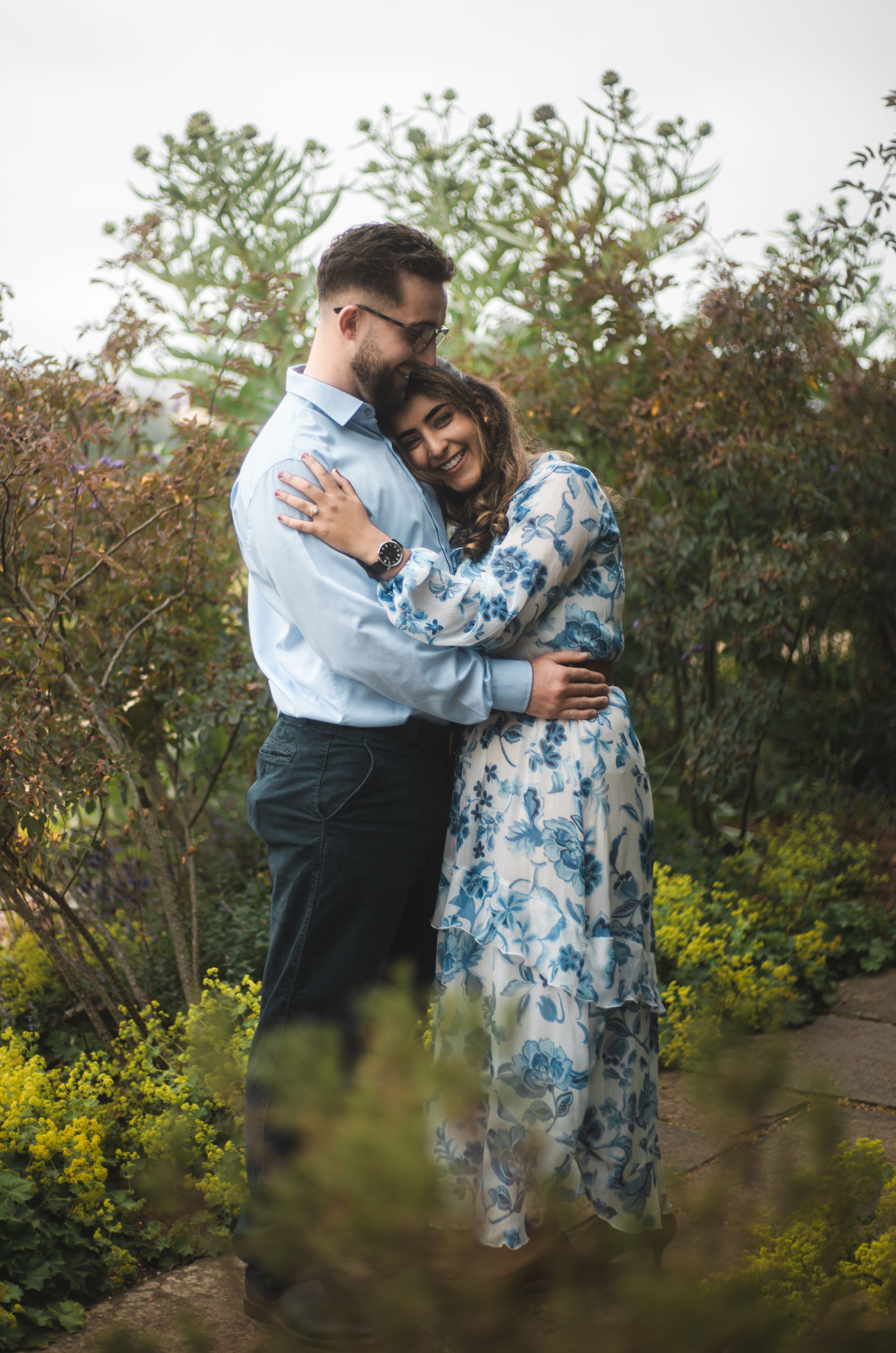 Couple embracing in a lush garden, woman in blue floral dress smiling, man in light blue shirt holding her close.