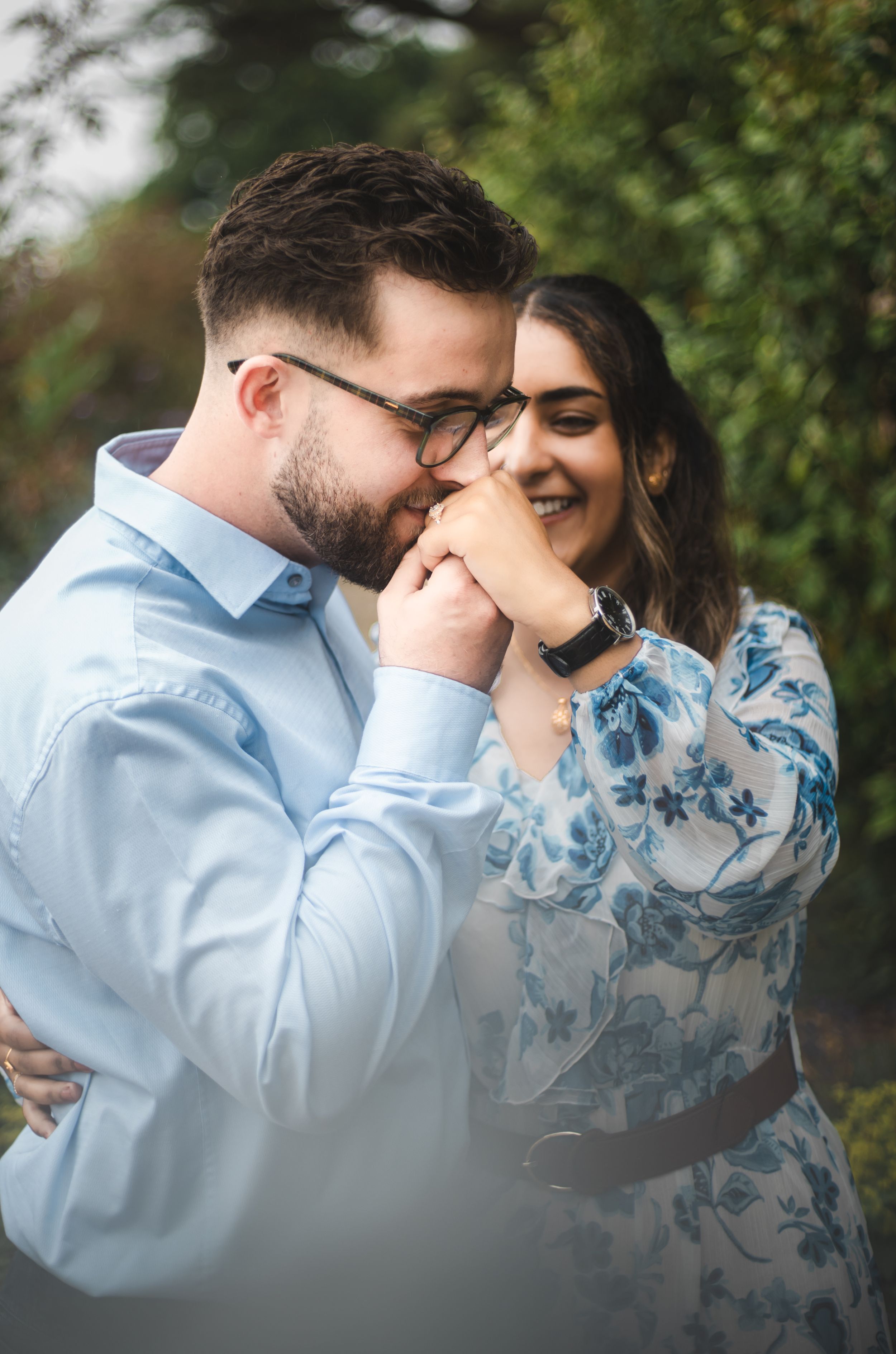 A couple shares a romantic moment outdoors, man in blue shirt kissing woman's hand as she smiles in floral dress.