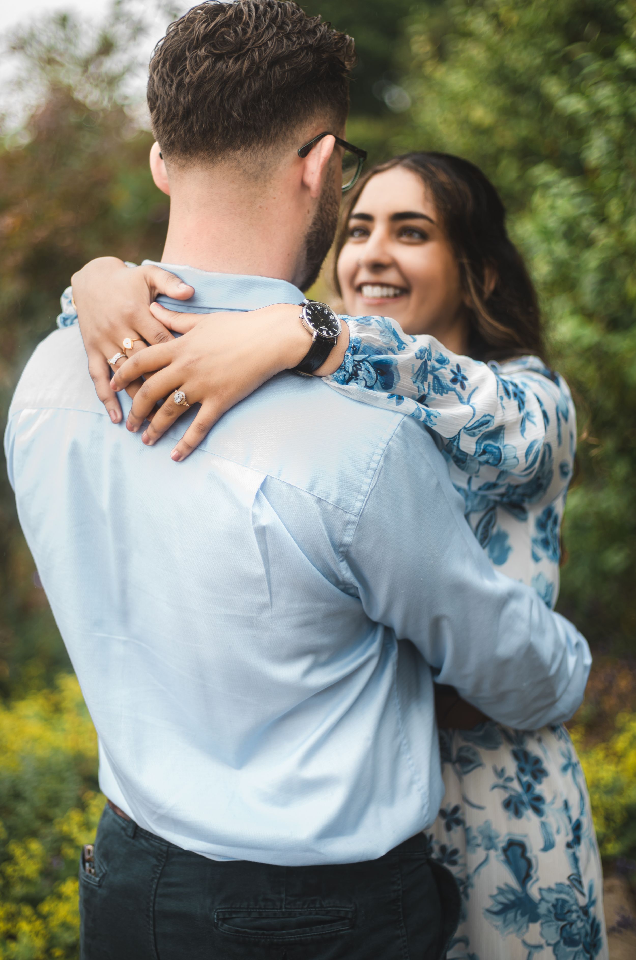 A smiling woman in a floral dress embraces a man in a light blue shirt outdoors among green foliage.