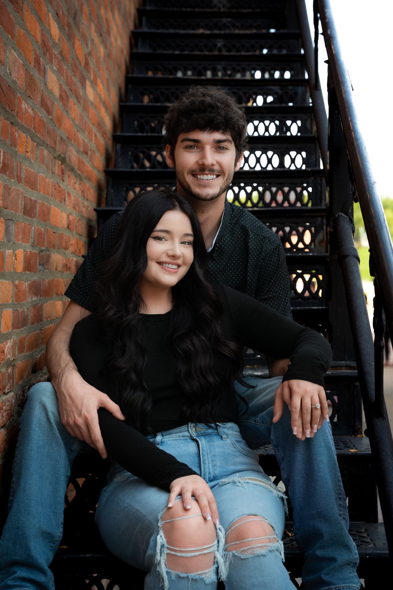 Happy couple sitting close together on outdoor metal staircase by brick wall.